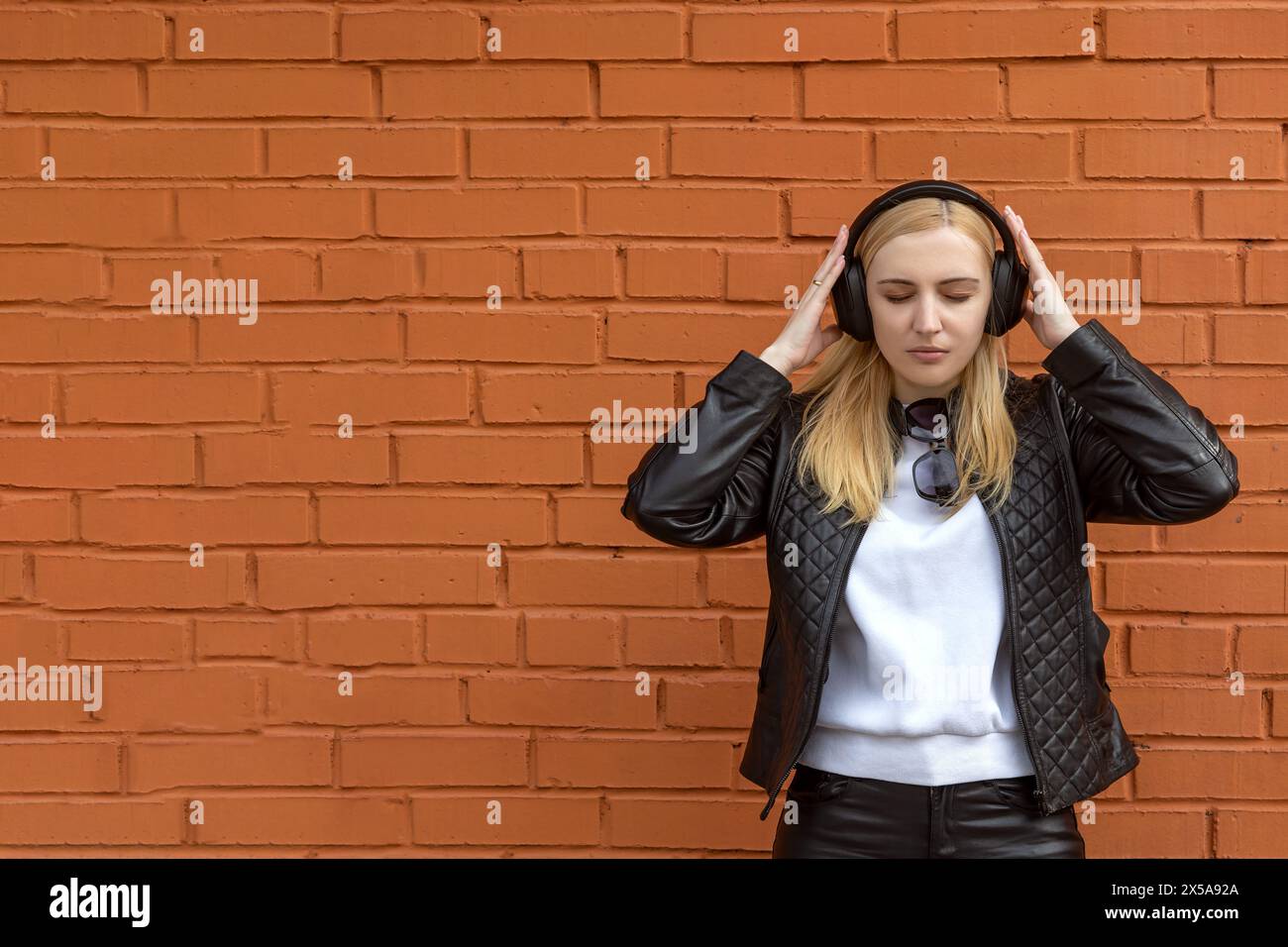 A stylish young woman is enjoying music on her headphones outdoors, standing against a vibrant orange brick wall backdrop Stock Photo