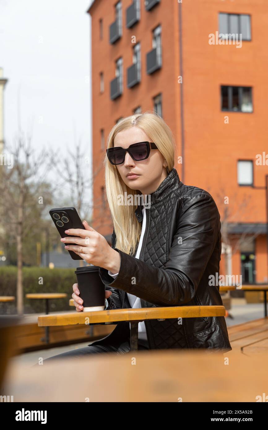 A fashionable woman wearing sunglasses and a black leather jacket is seated with a coffee, absorbed in her smartphone on an urban street with a brick Stock Photo