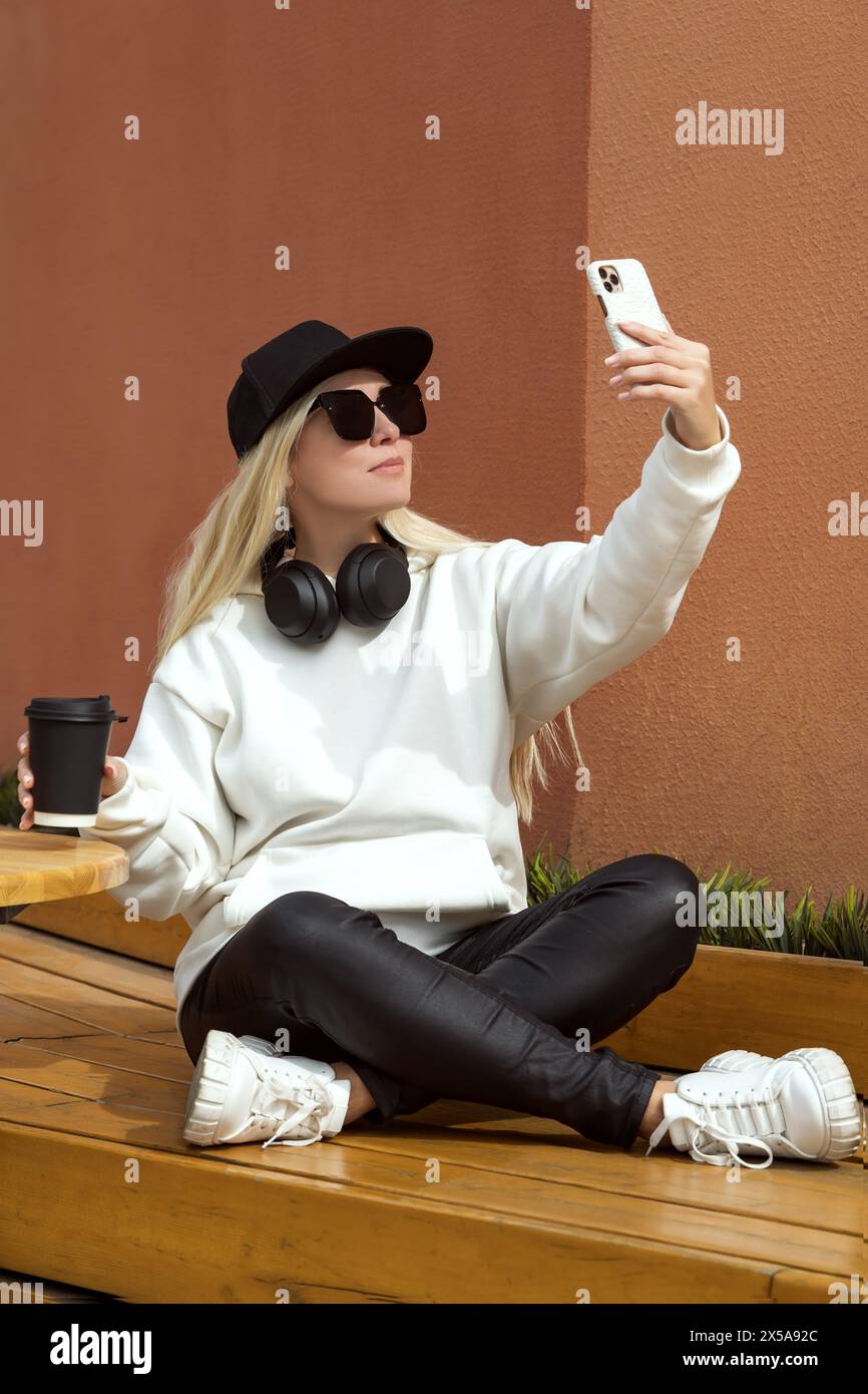Fashionable blonde woman in casual wear, sitting on a bench and taking a selfie with a mobile phone in an urban setting. Stock Photo