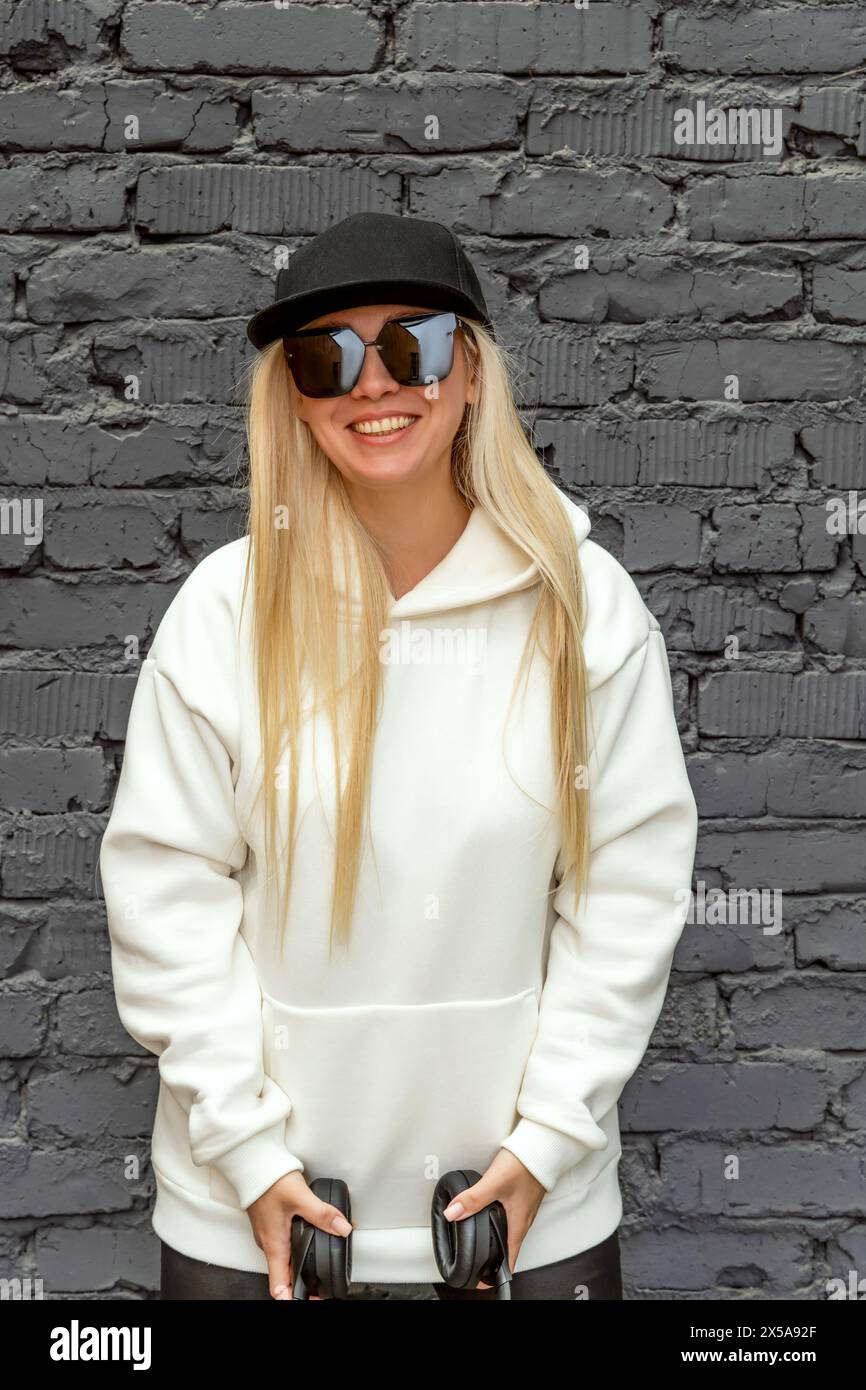 Blonde woman smiles on a city street, wearing a cap, sunglasses, and holding headphones. Stock Photo