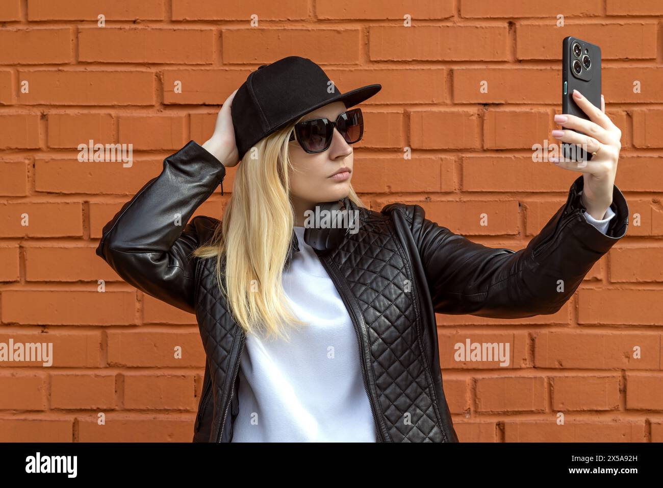 Fashionable young woman in a black leather jacket and hat taking a selfie with her smartphone against an orange brick wall background Stock Photo