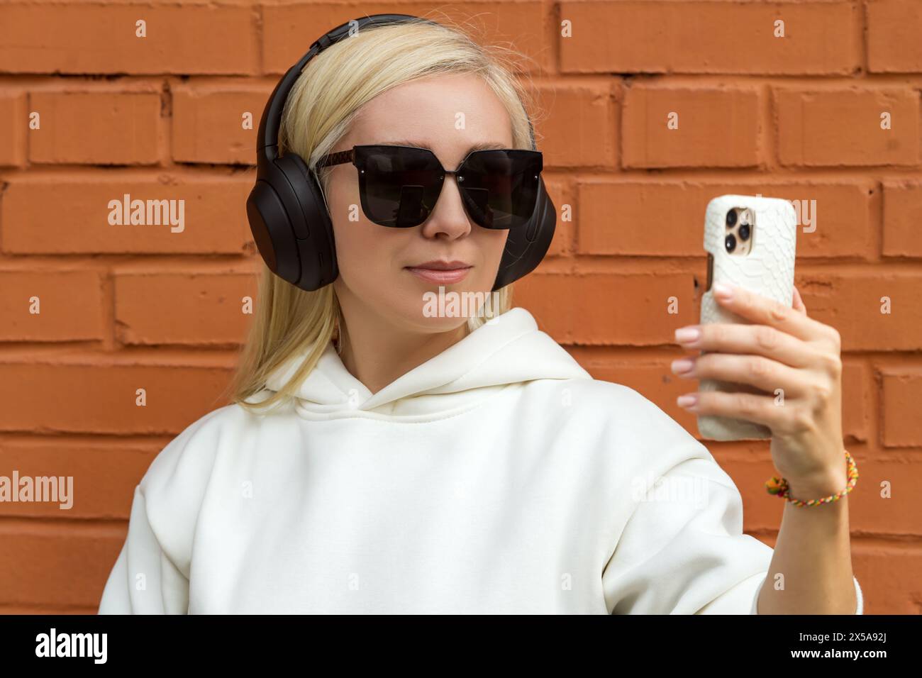 A stylish blonde woman wearing large sunglasses and headphones holds a smartphone, seemingly taking a selfie against a brick wall backdrop. Stock Photo