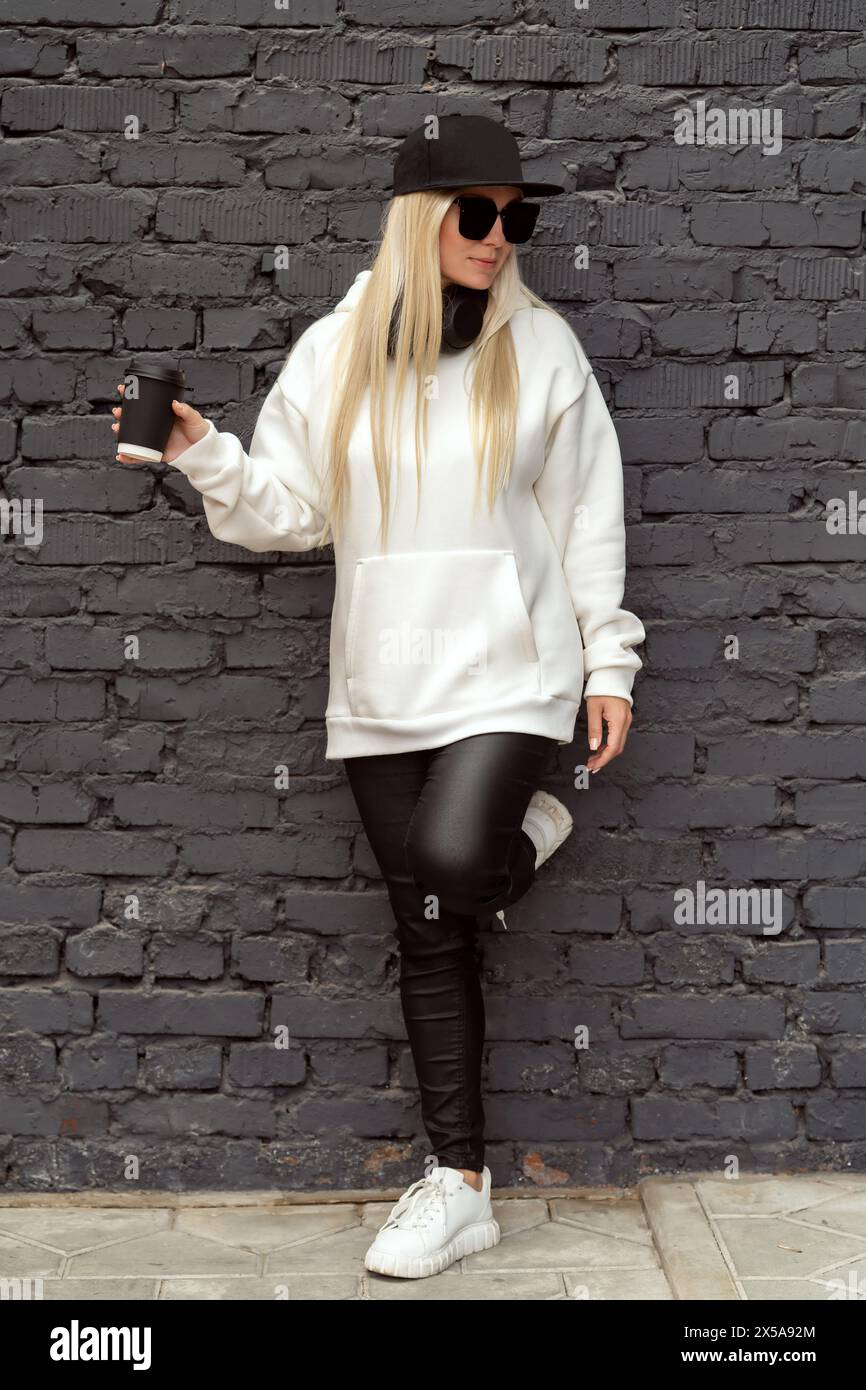 A cool blonde woman in casual attire enjoys her coffee against a textured brick wall. Stock Photo