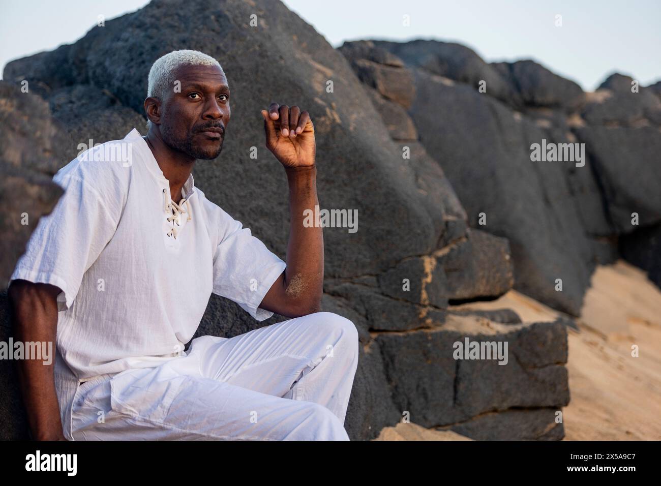 An elegant African American male model poses on a tranquil beach, dressed in white, exuding a peaceful aura against a backdrop of rocks Stock Photo