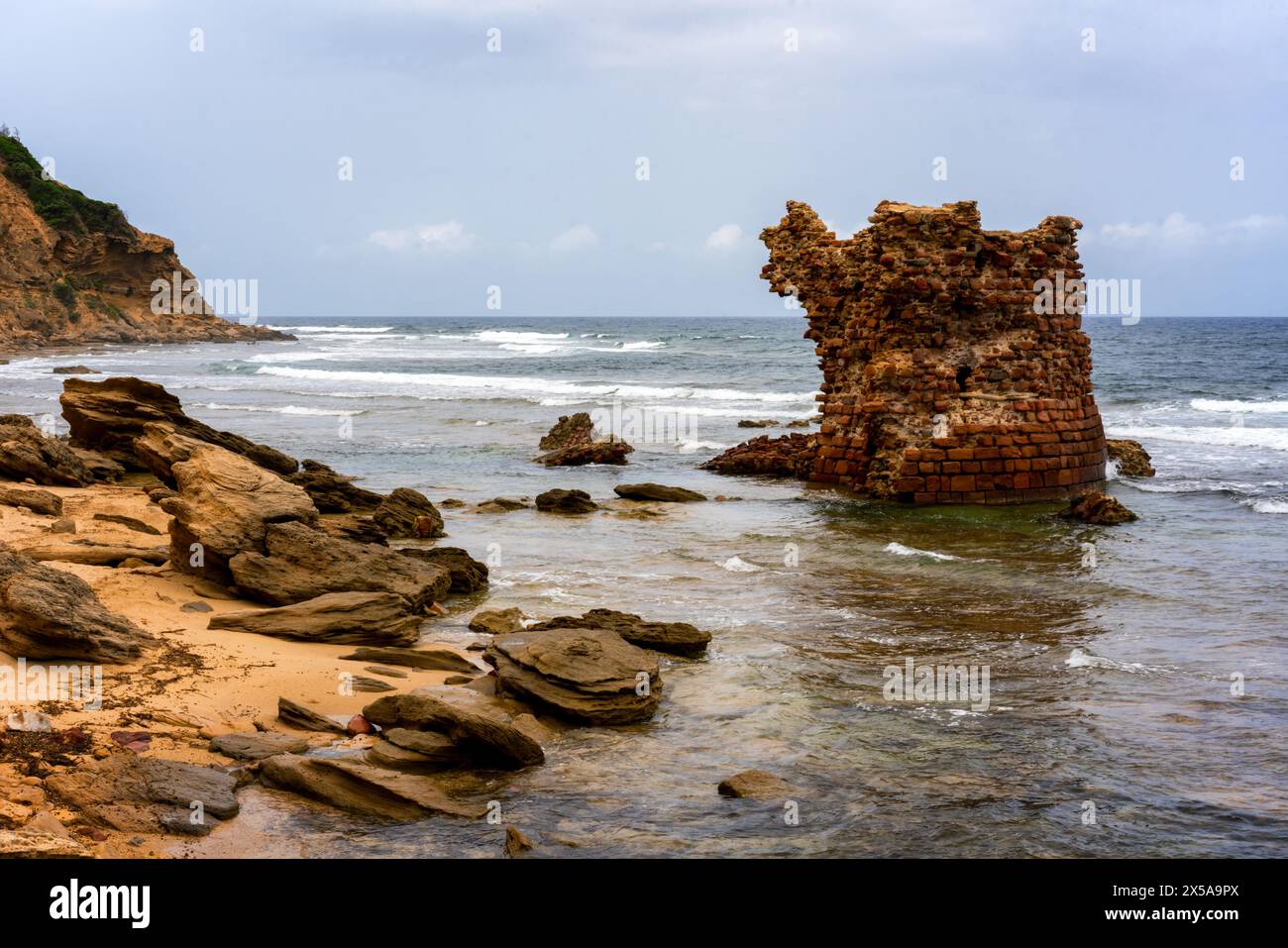 Ruins of a historic tower standing on a rocky Porto Paglia beach under a cloudy sky, symbolizing past civilizations Stock Photo