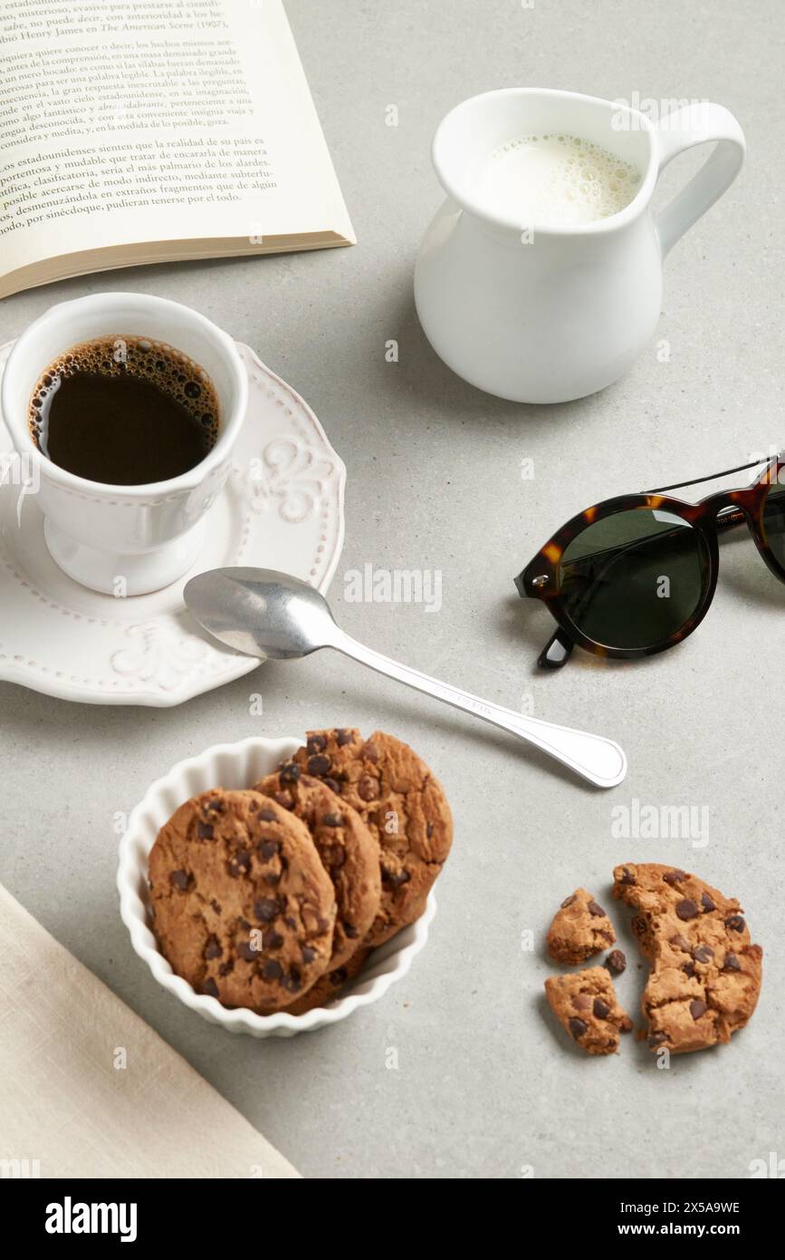 A cozy tabletop view featuring a cup of coffee, cookies on a plate, and a milk jug, accompanied by reading glasses and an open book Stock Photo