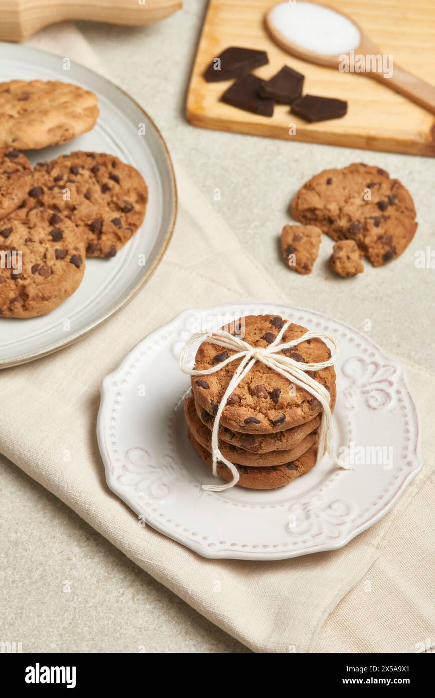 A charming presentation of chocolate chip cookies, tied with twine on a decorative white plate, with more cookies and ingredients nearby Stock Photo