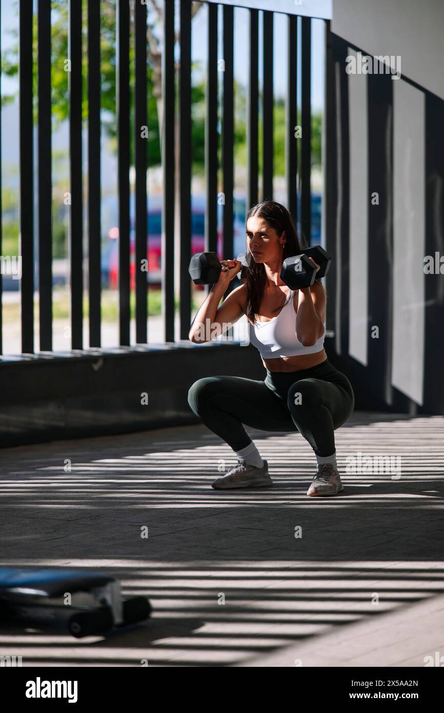 A focused sportswoman performs a squat with dumbbells at home, showcasing a dedication to her fitness regimen Stock Photo