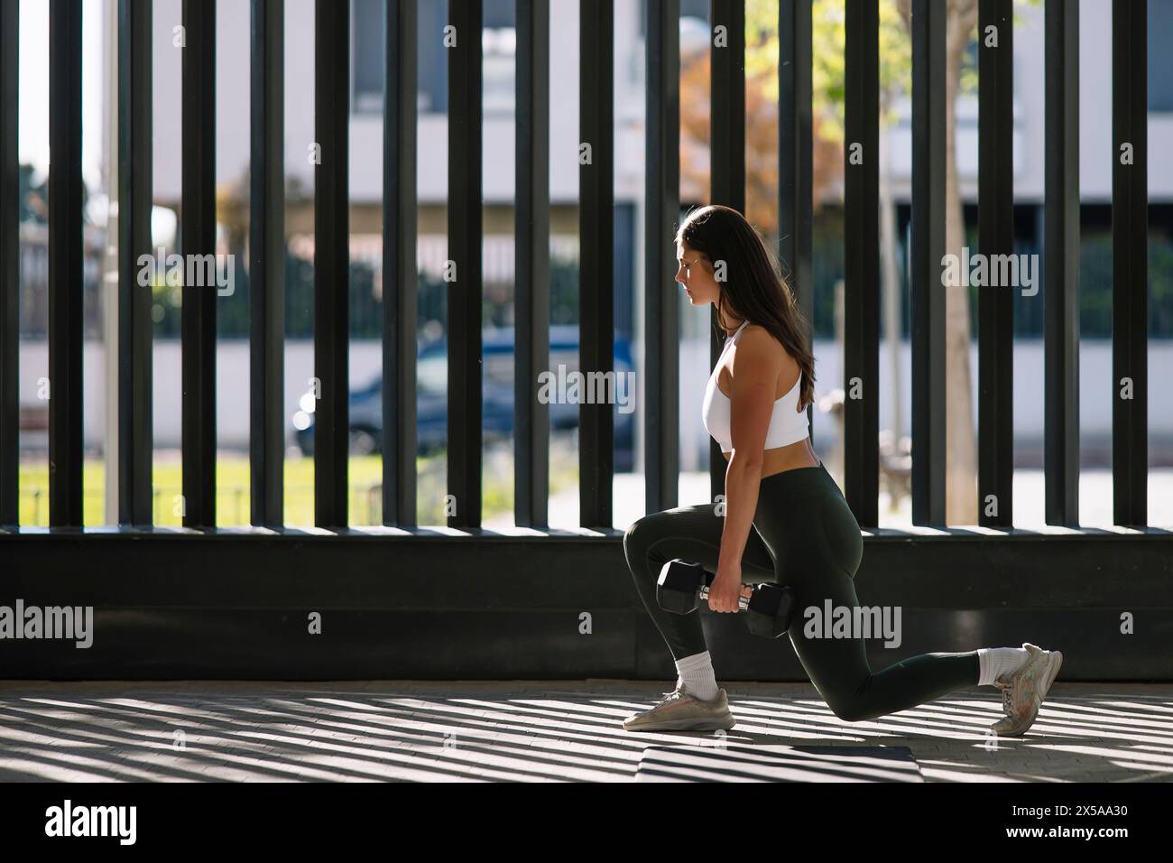 A focused young woman in sportswear performs lunges with a dumbbell in a bright, sunlit home environment, depicting a healthy lifestyle Stock Photo
