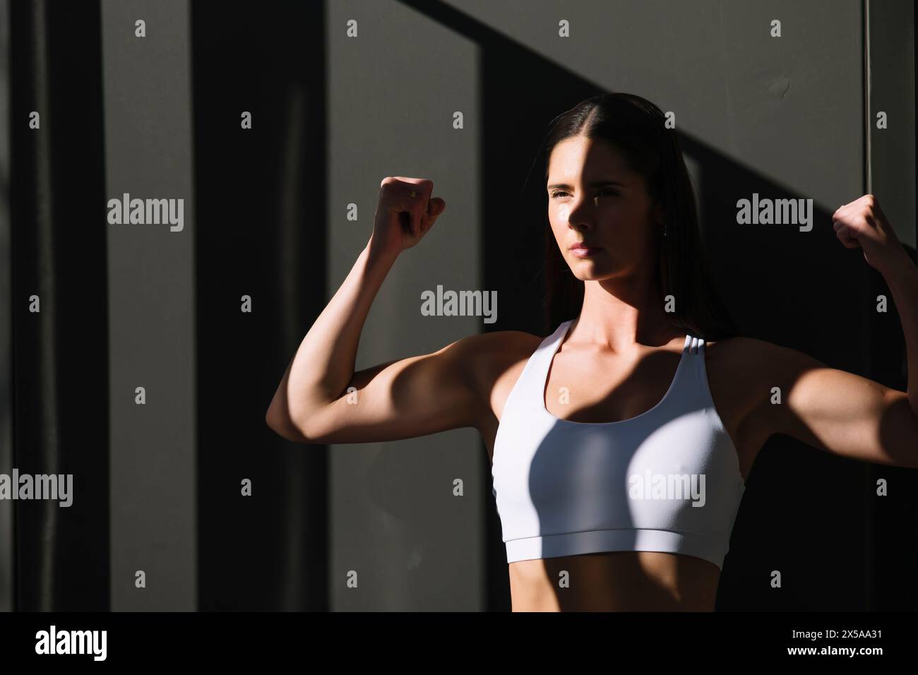 A fit woman in sportswear flexes her muscles confidently, illuminated by natural sunlight with a shadowy background Stock Photo
