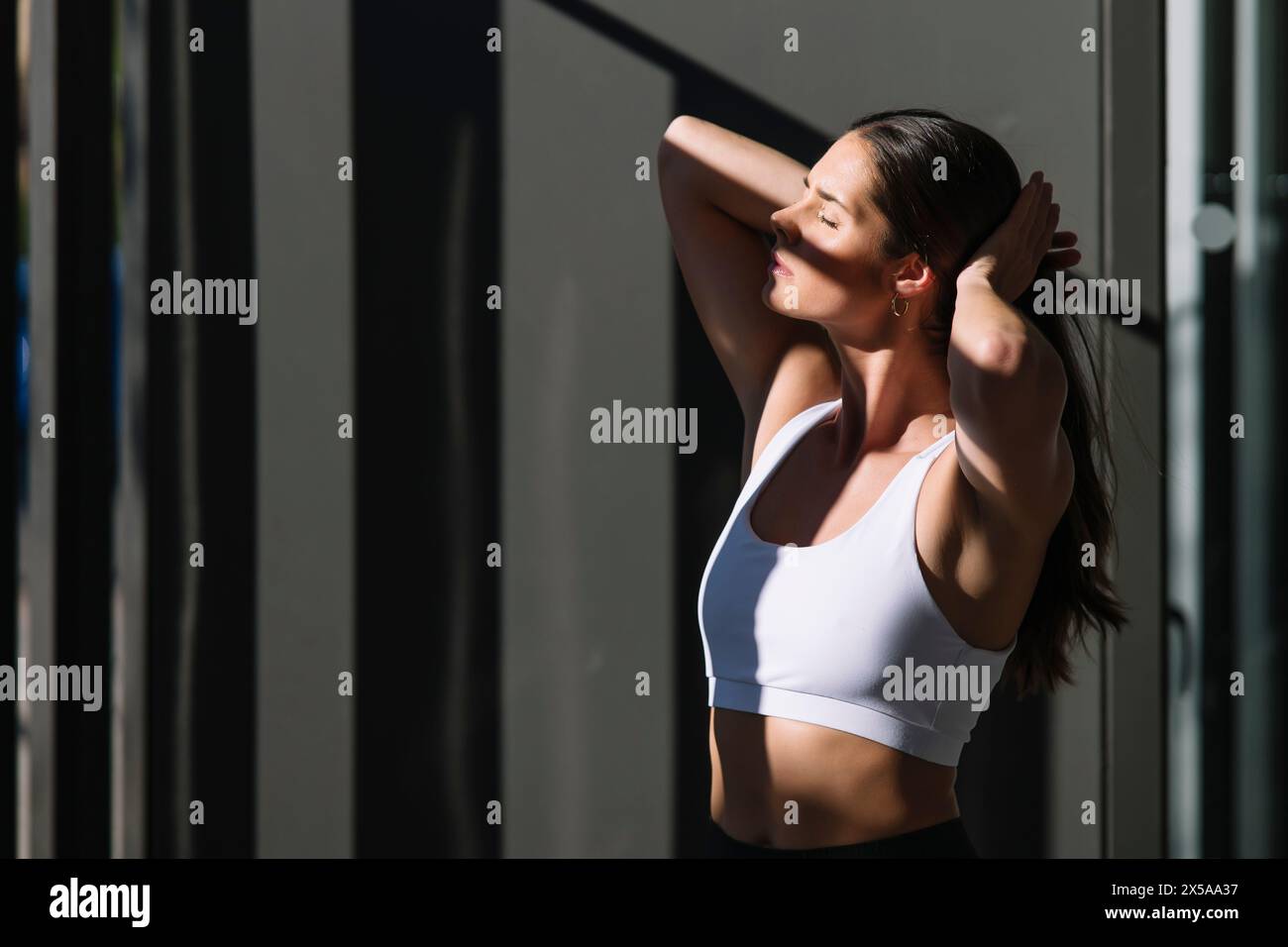 A fit woman in sportswear performs a stretch at home, basking in the sunlight that enhances her peaceful expression Stock Photo