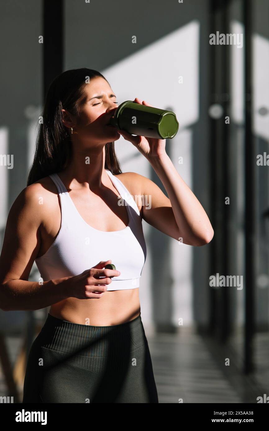 A fit woman hydrates by drinking water from a green bottle, post-exercise, in a well-lit home setting Stock Photo