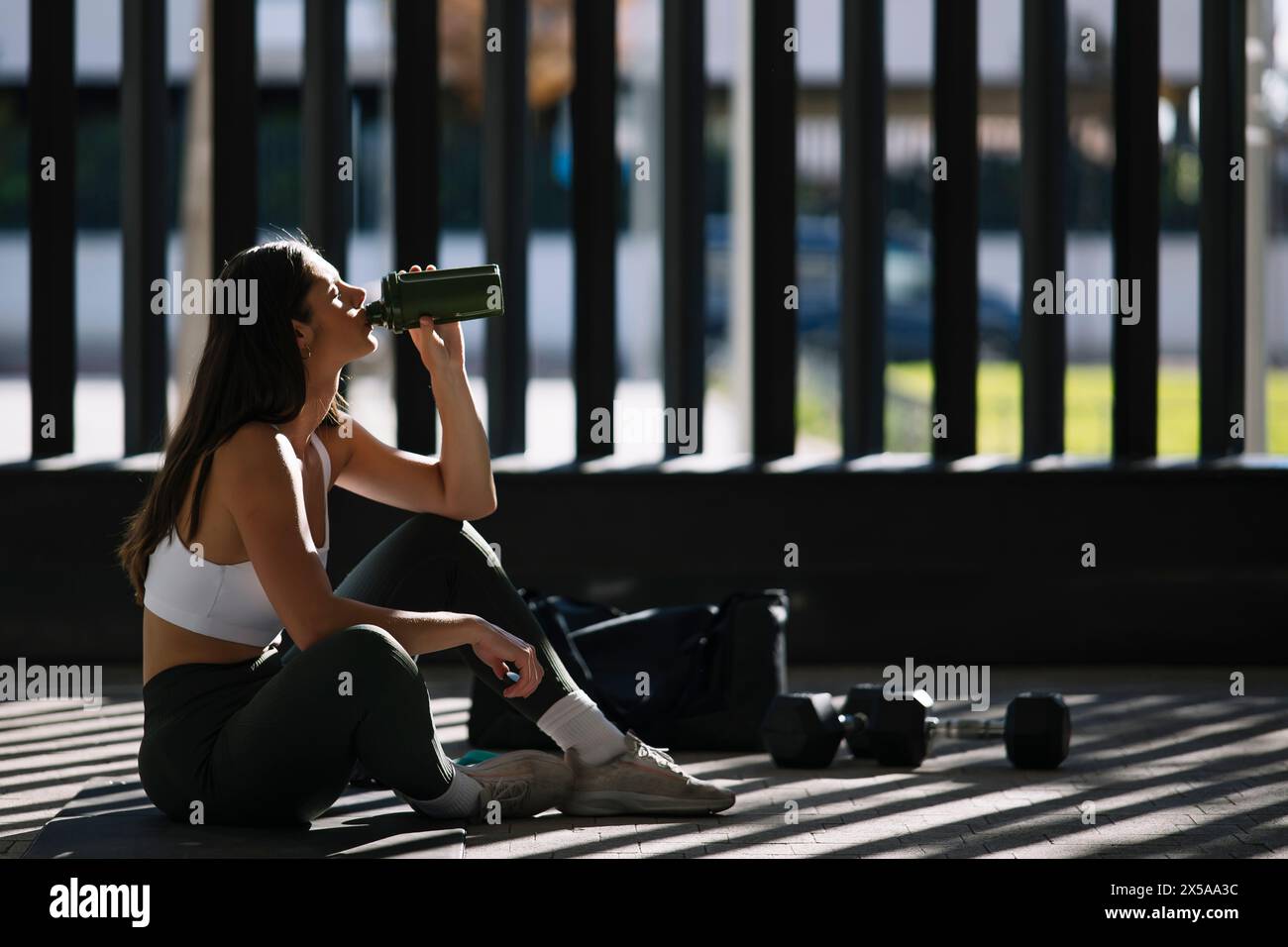 An athletic woman takes a break to drink water in a home gym setting, with sunlight streaming through windows and exercise equipment nearby Stock Photo