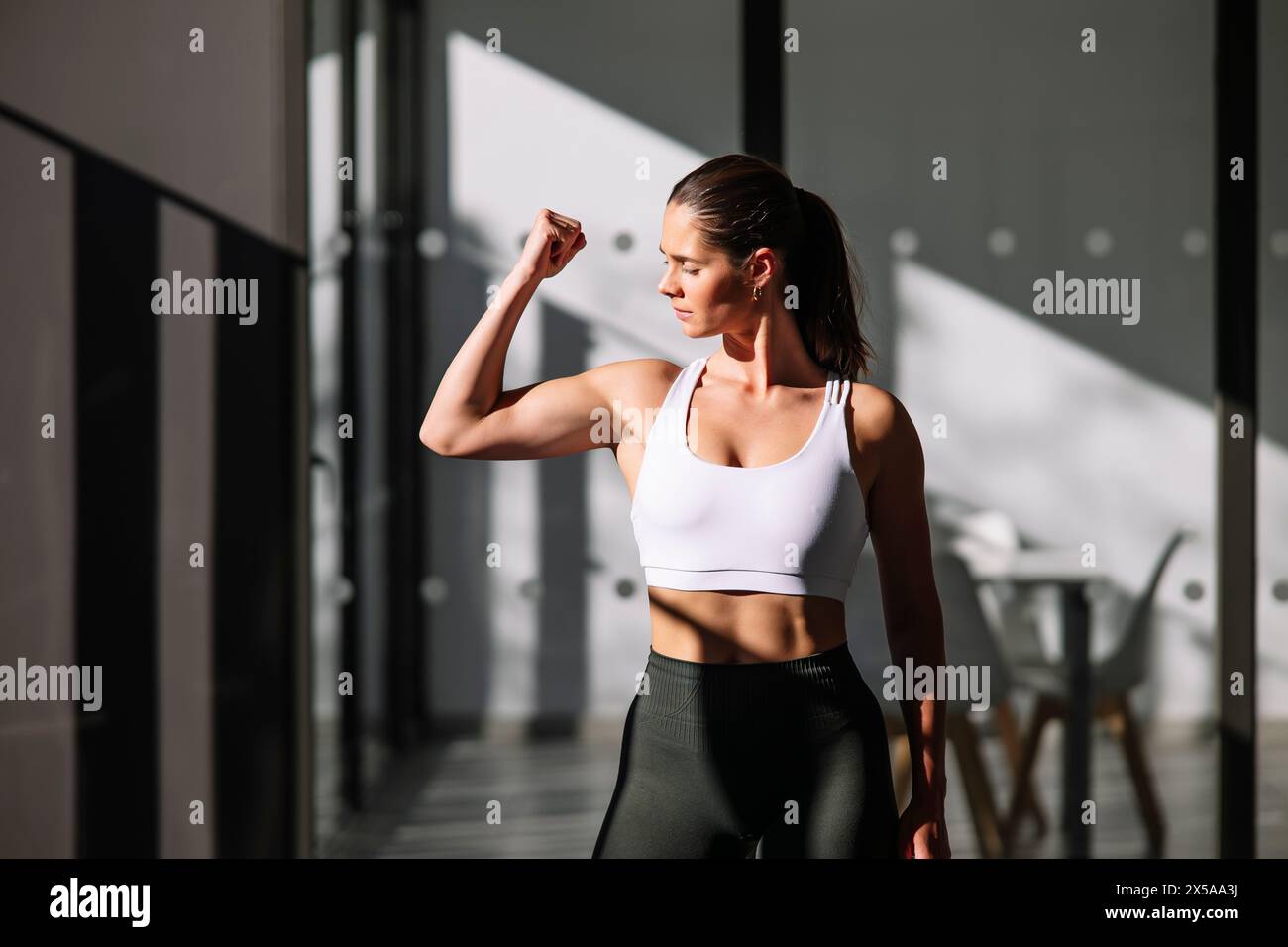 A physically fit woman in sportswear flexes her muscles while exercising at home, showcasing strength and wellness Stock Photo