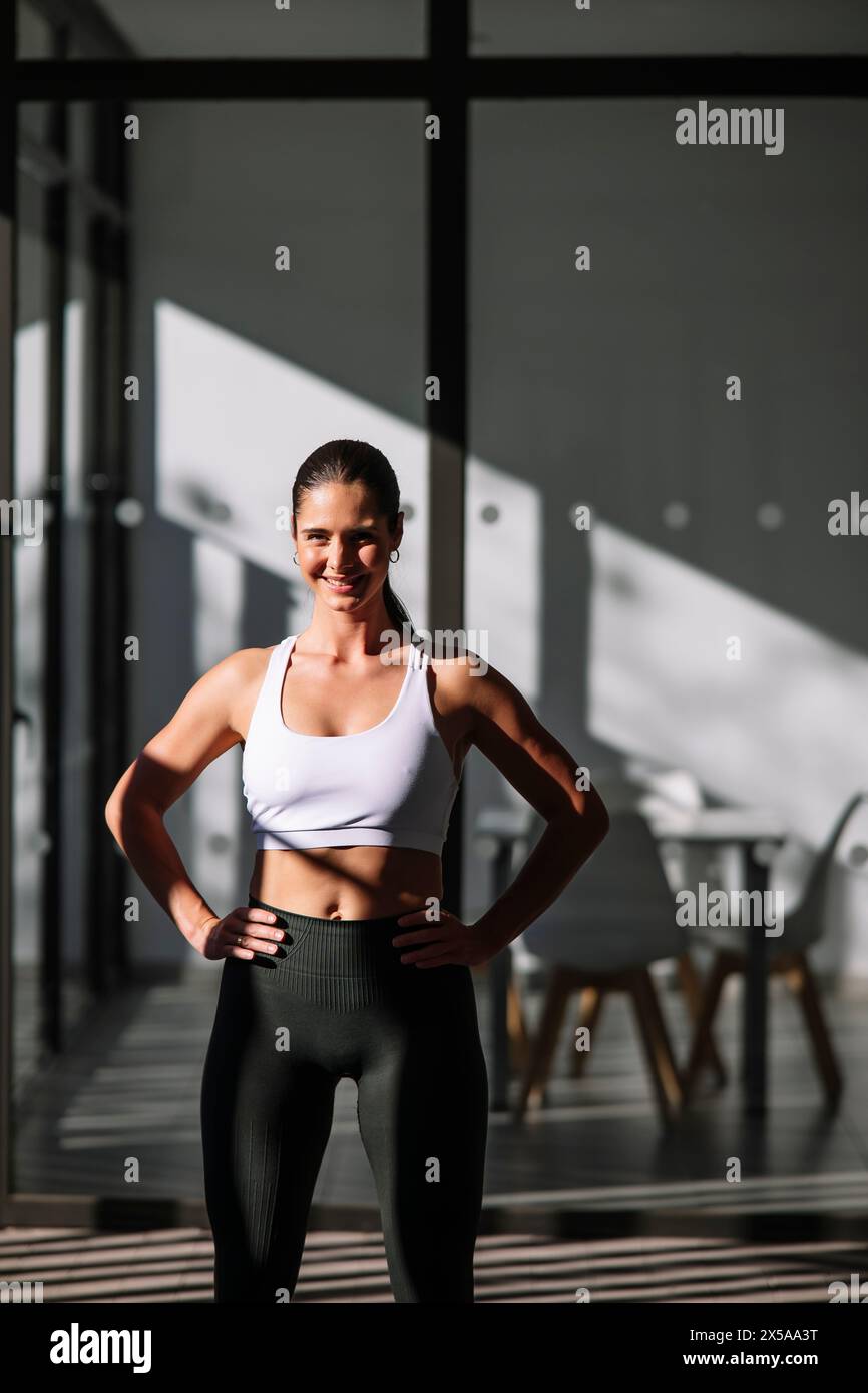 A fit woman with a beaming smile stands proudly in her home, showcasing her athletic physique in sporty attire against a backdrop of stylish home inte Stock Photo