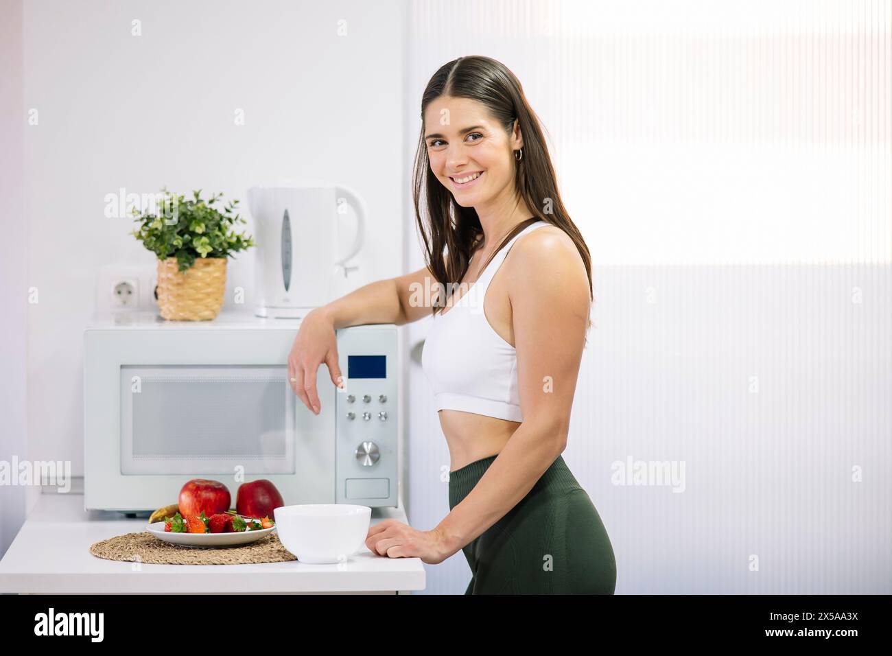 A fit woman in athletic wear smiles while preparing a healthy meal with fresh fruits in her modern home kitchen Stock Photo
