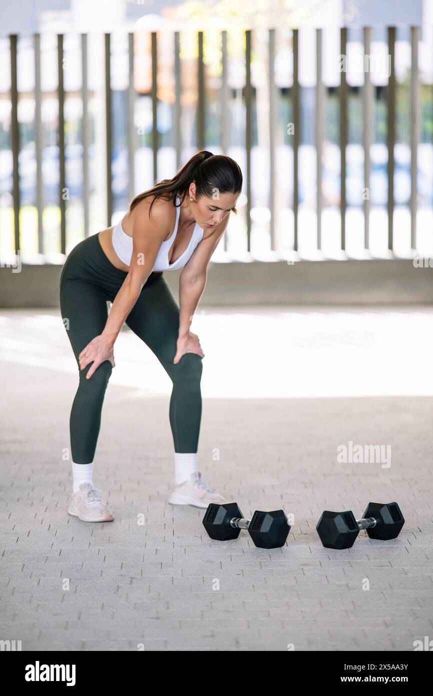 Fit woman pauses, hands on knees, after exercising with dumbbells at home, showcasing an active lifestyle and indoor fitness routine Stock Photo