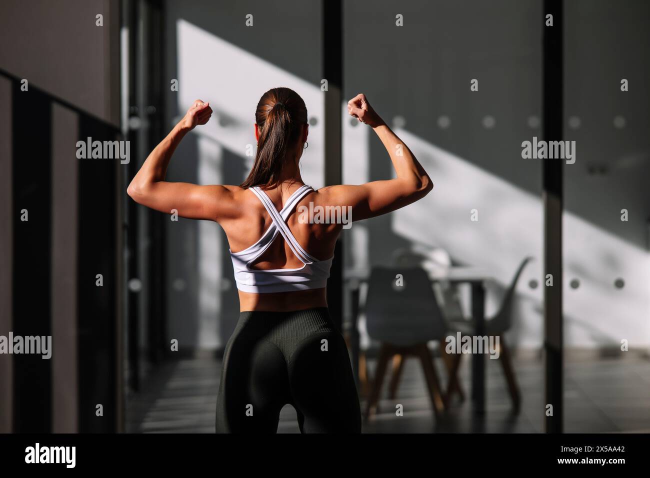 A strong female athlete showcases her toned back muscles while flexing in a modern home environment, displaying fitness and strength Stock Photo