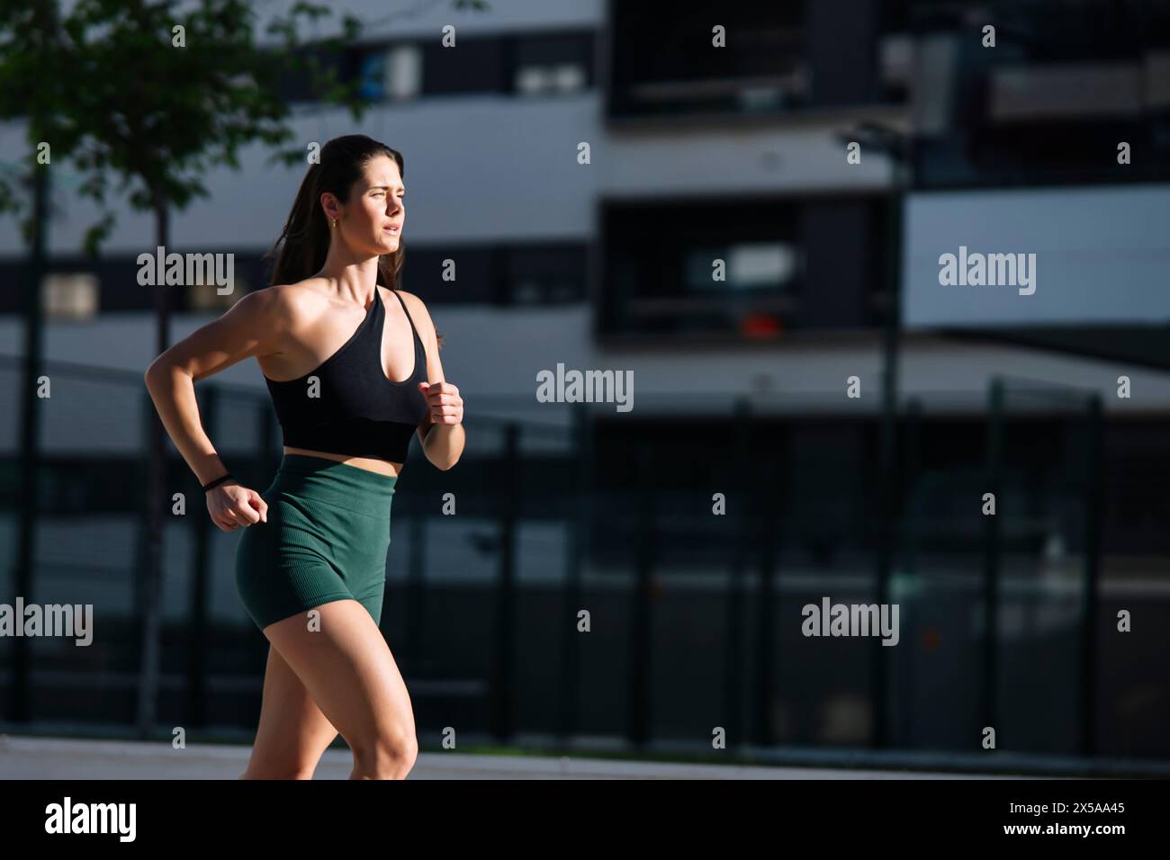 An athletic woman in sports attire jogs against a backdrop of modern buildings, showcasing a healthy lifestyle in an urban environment Stock Photo