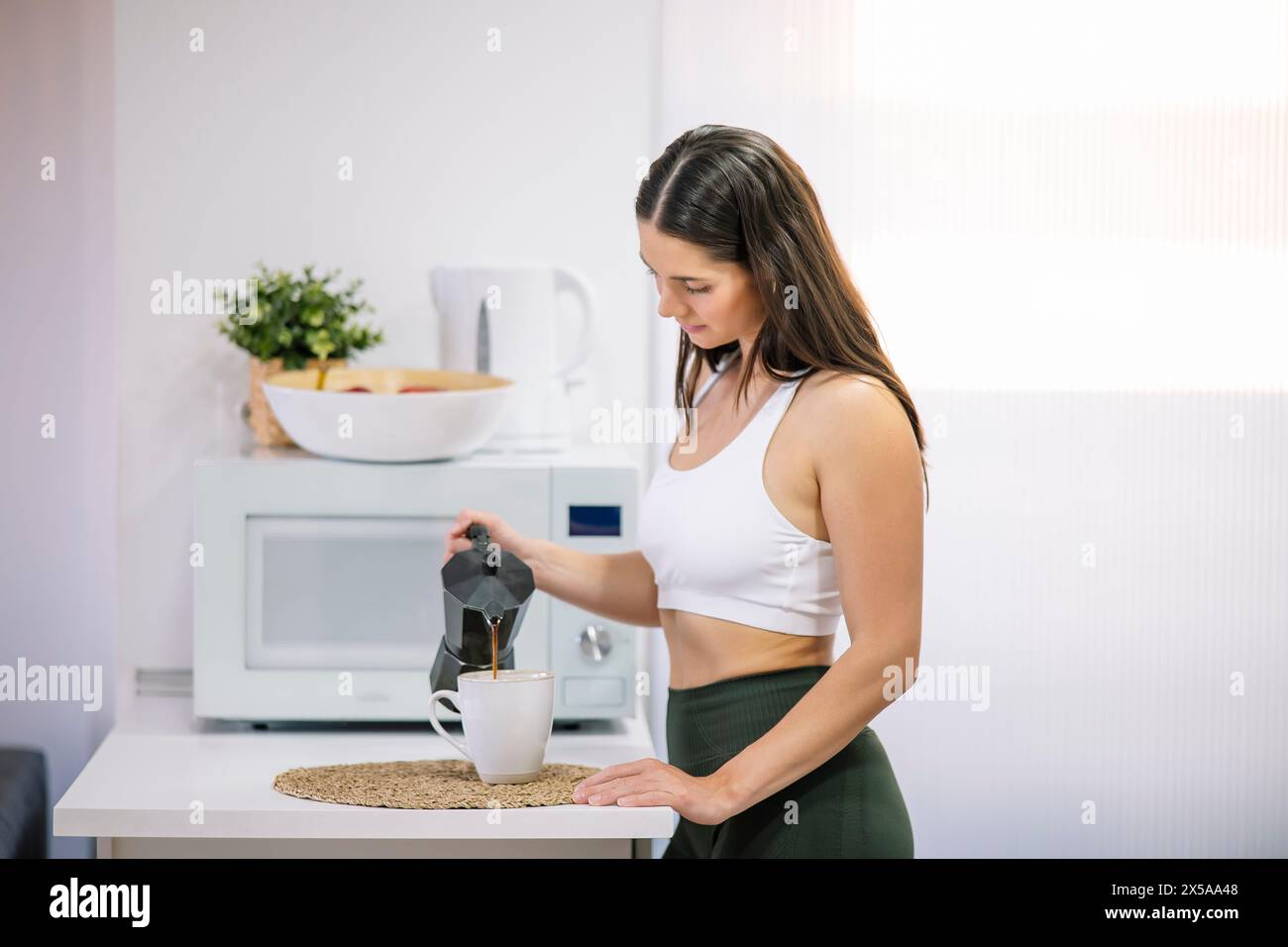 A fit woman in athletic wear pours coffee from a moka pot into a mug in her well-lit home kitchen, exemplifying a healthy lifestyle Stock Photo