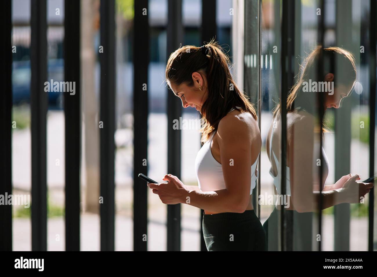 An athletic woman pauses her workout at home to check her smartphone, standing by a window with sunlight filtering through Stock Photo