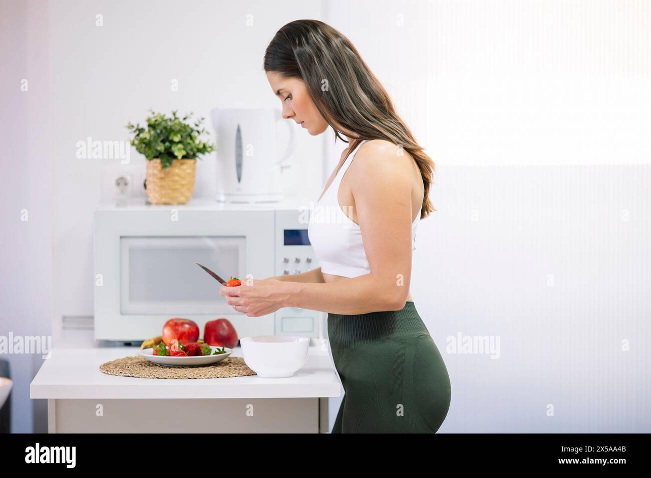 A fitness-focused woman in activewear is slicing vegetables for a nutritious meal in her modern kitchen, exemplifying a healthy lifestyle Stock Photo