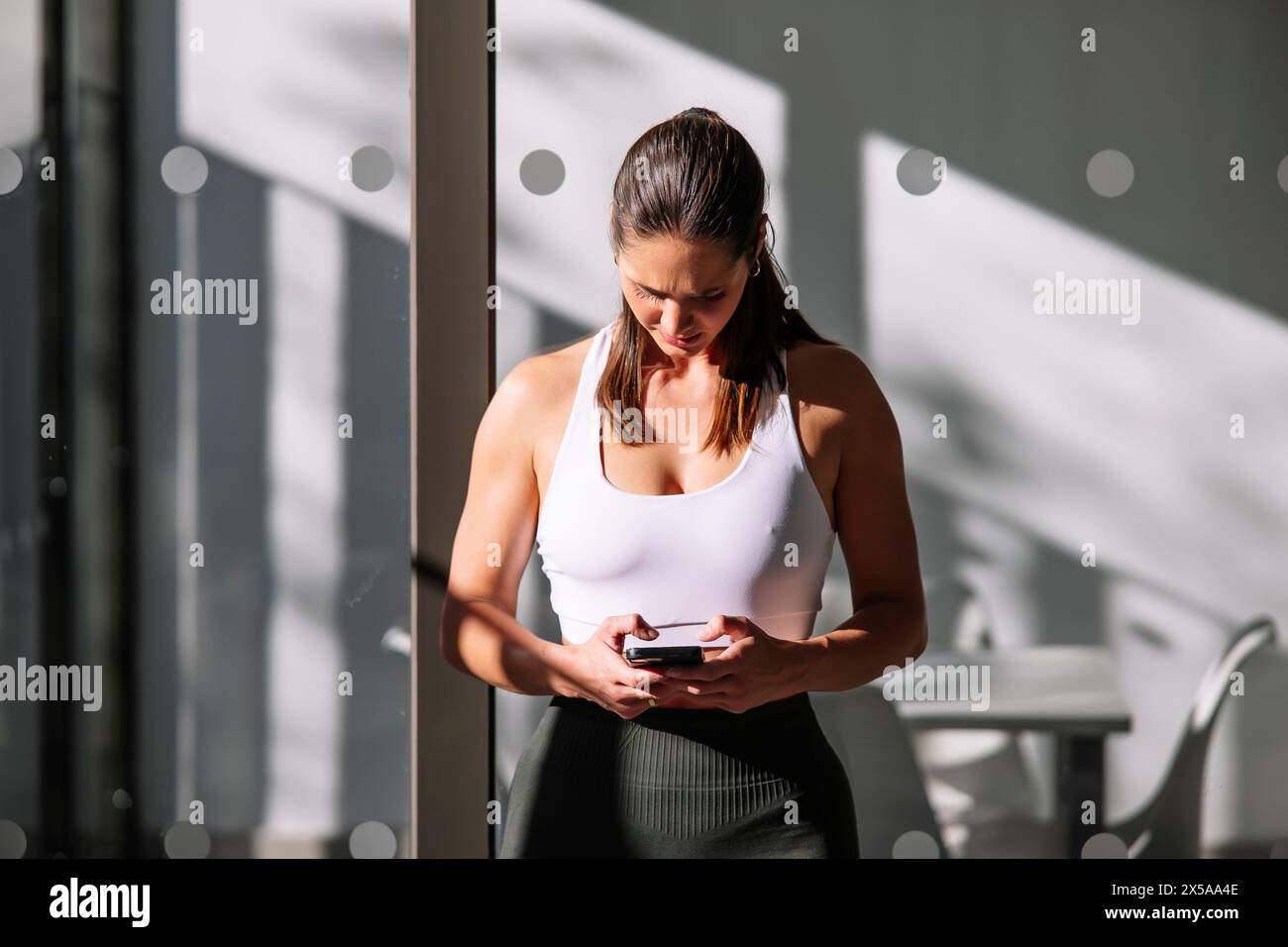 A focused athletic woman checks her smartphone in a sunlit room, showcasing a healthy lifestyle at home Stock Photo