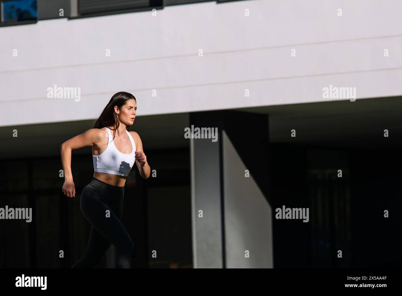 An athletic woman is captured mid-run, sporting a white sports bra and black leggings, showcasing an active lifestyle amidst an urban backdrop Stock Photo