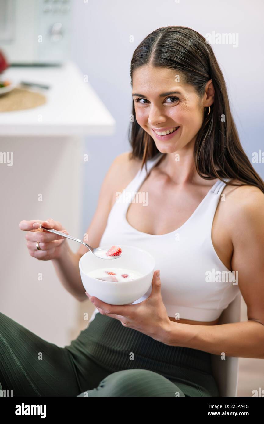 A fit woman in sportswear smiles while eating a bowl of yogurt with strawberries in a modern kitchen, representing a healthy lifestyle Stock Photo