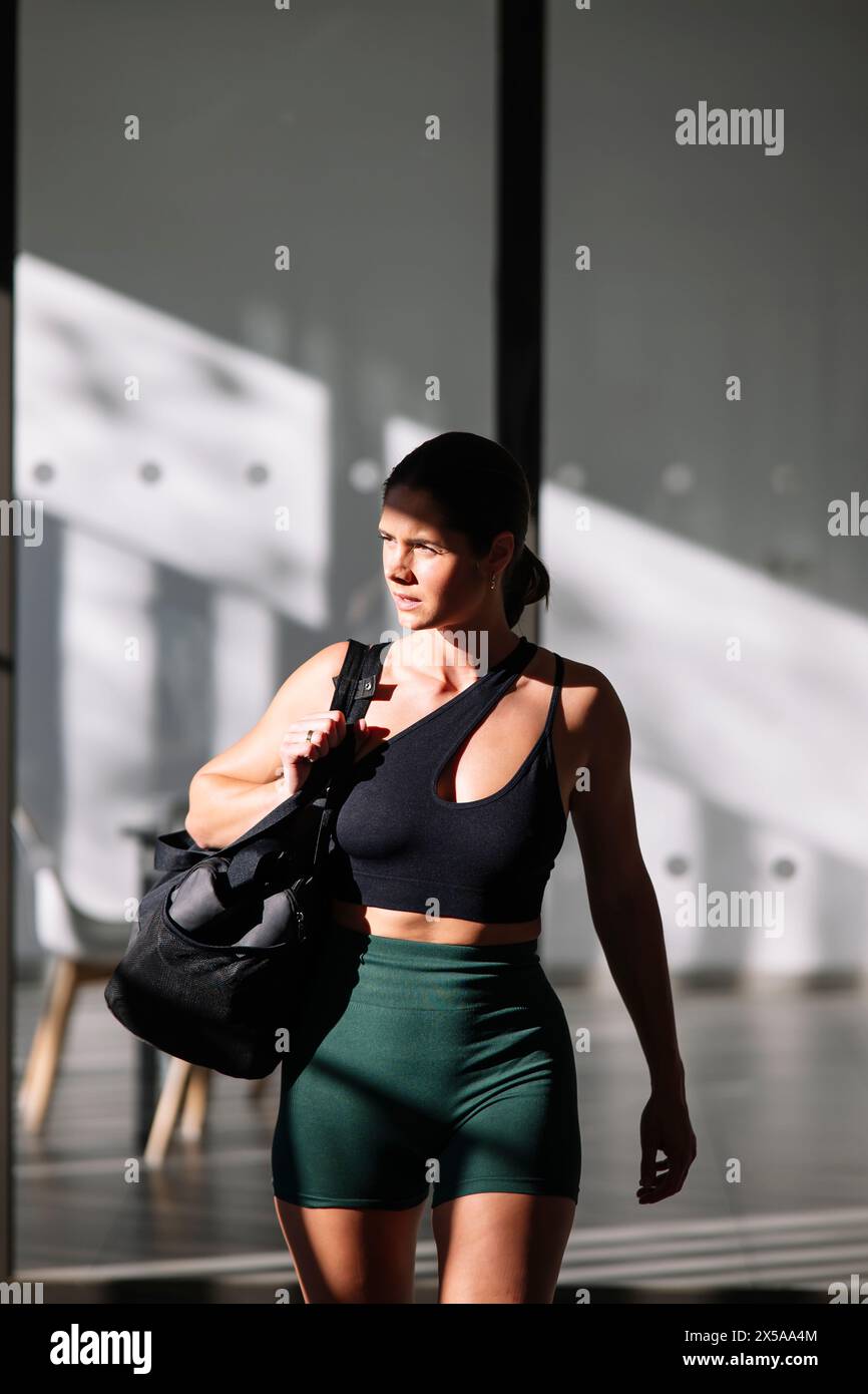 Athletic woman in sportswear holding a gym bag, standing in a well-lit home with a modern interior, preparing for a fitness session Stock Photo