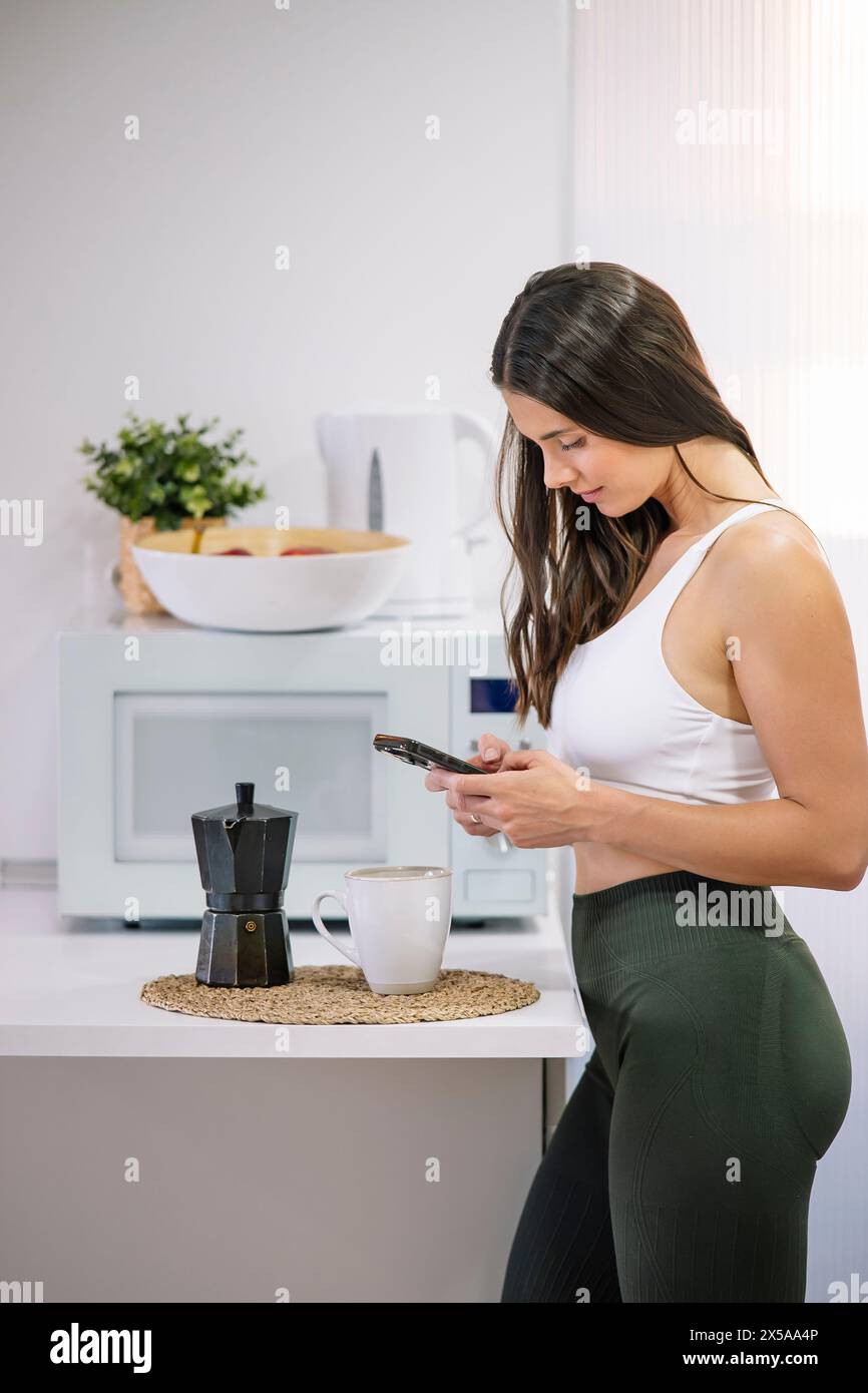 A fit woman in sportswear engages with her smartphone in a modern kitchen setting with a coffee maker and mug on the counter Stock Photo