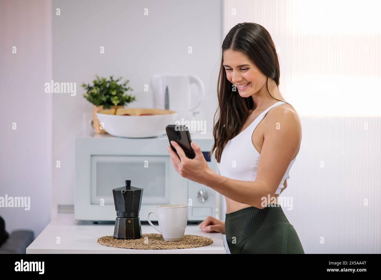 A fit woman in activewear smiles while using her smartphone in a modern kitchen Stock Photo