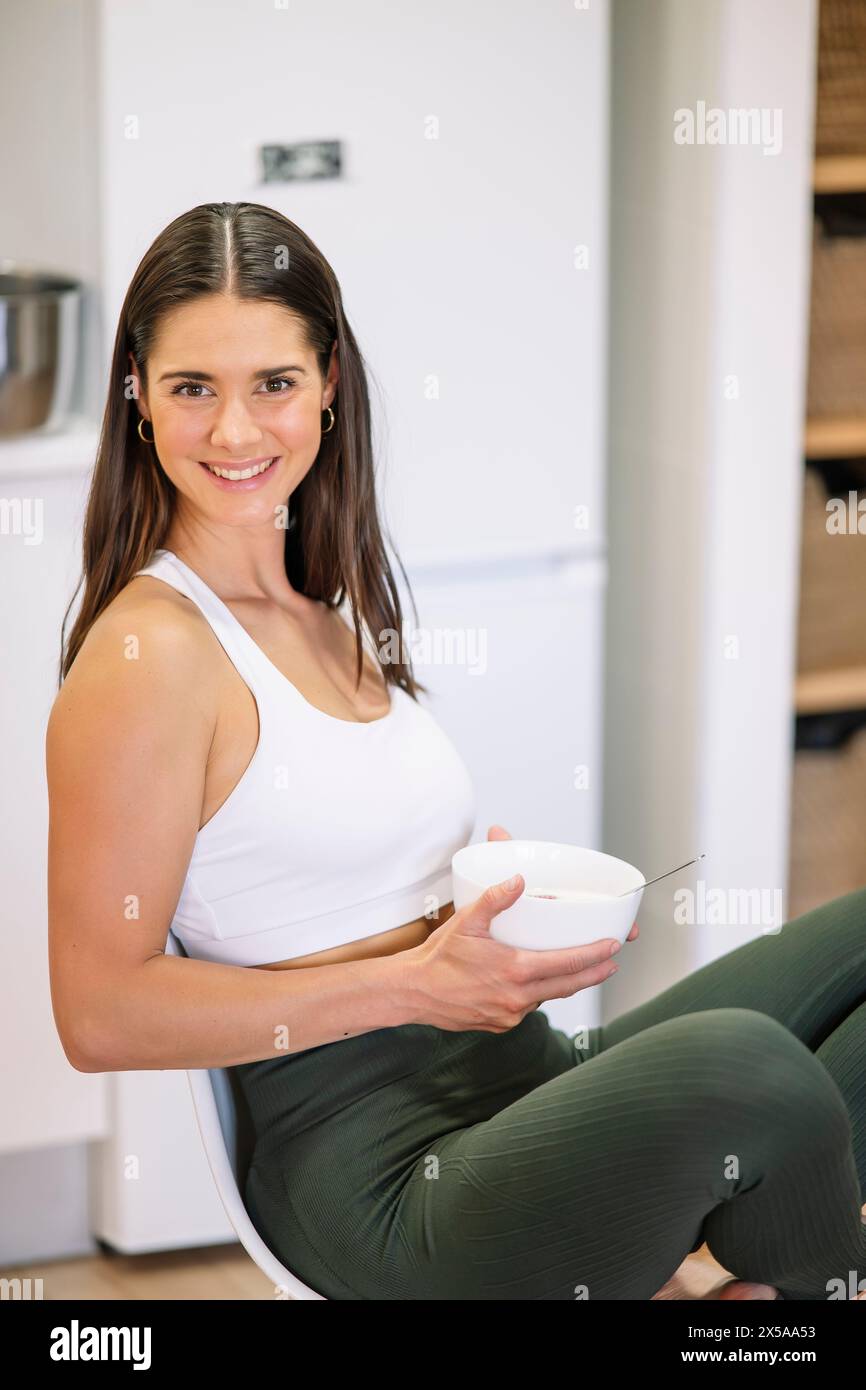 A cheerful athletic woman in sportswear enjoys a healthy meal in a modern kitchen setting Stock Photo