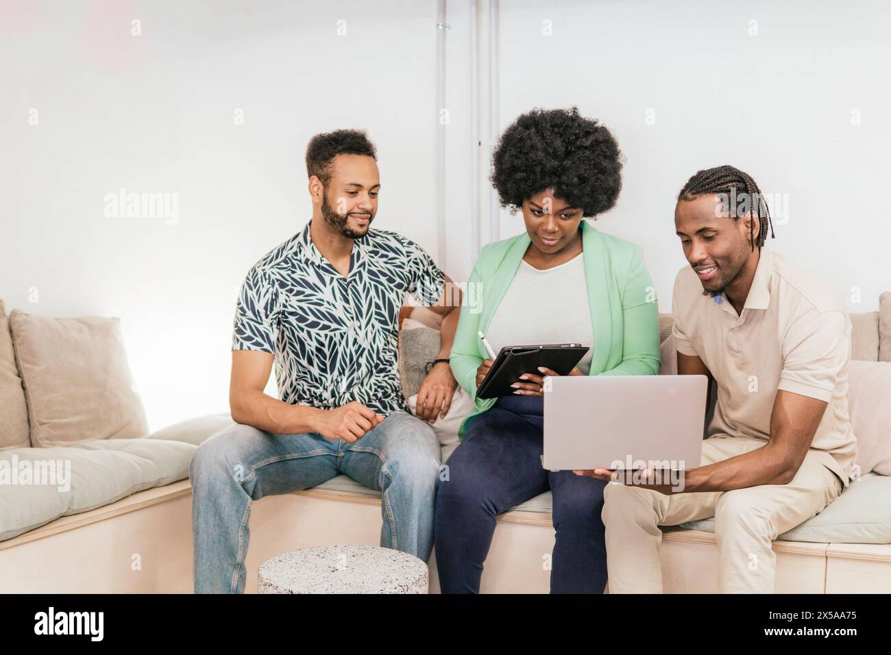 Three professionals engage in a coworking session, sharing ideas on a tablet and a laptop in a comfortable home environment Stock Photo