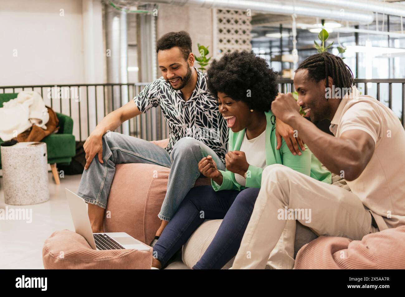 Three colleagues cheer in excitement in a modern coworking space while looking at the laptop screen Stock Photo