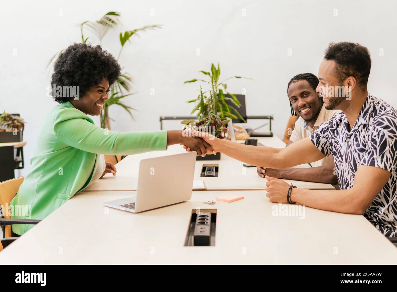 Three professionals happily engaging in a fist bump over a work table in a bright coworking environment Stock Photo