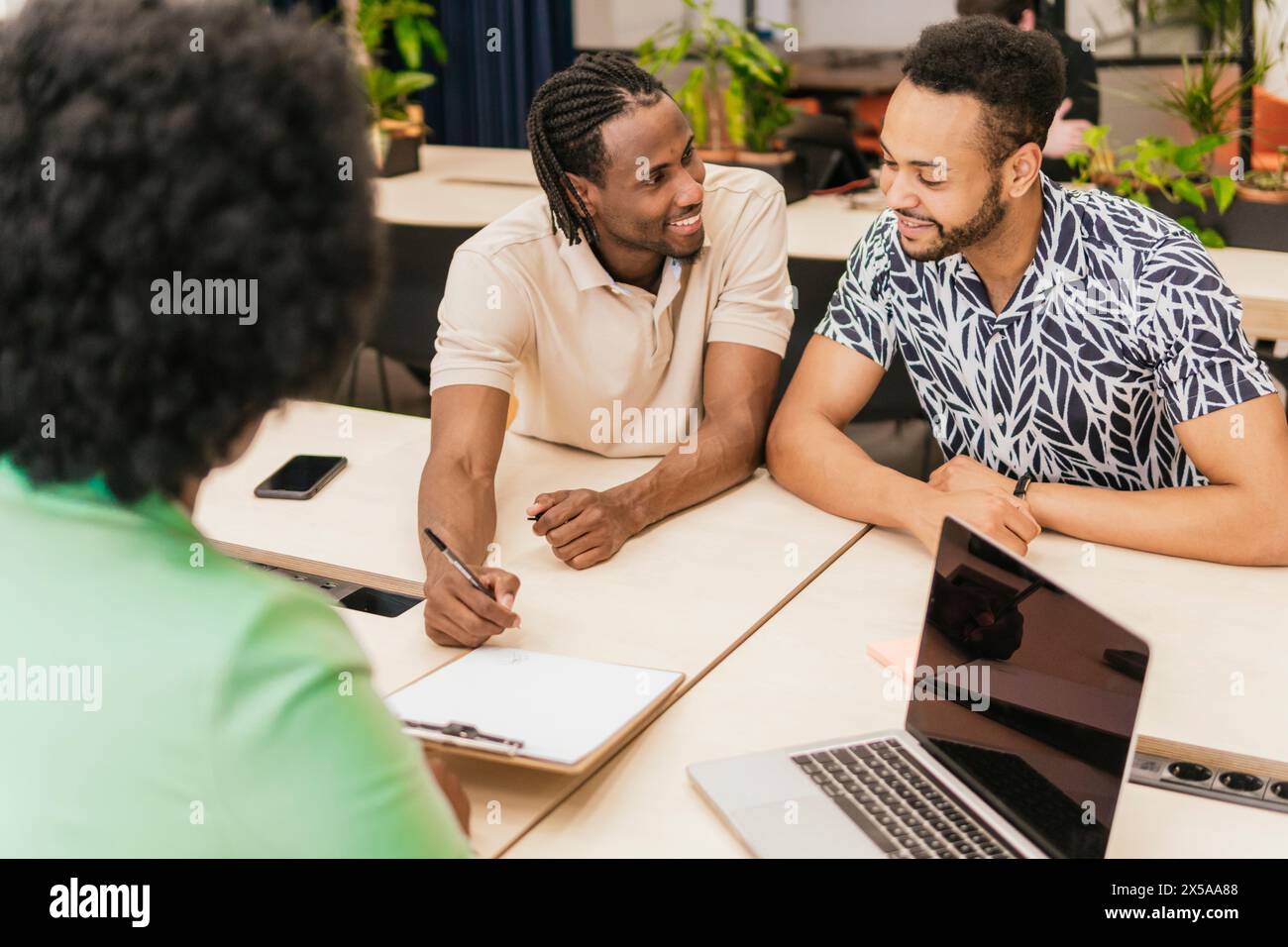 Three professionals collaborate warmly at a coworking space, sharing ideas and using digital devices Stock Photo