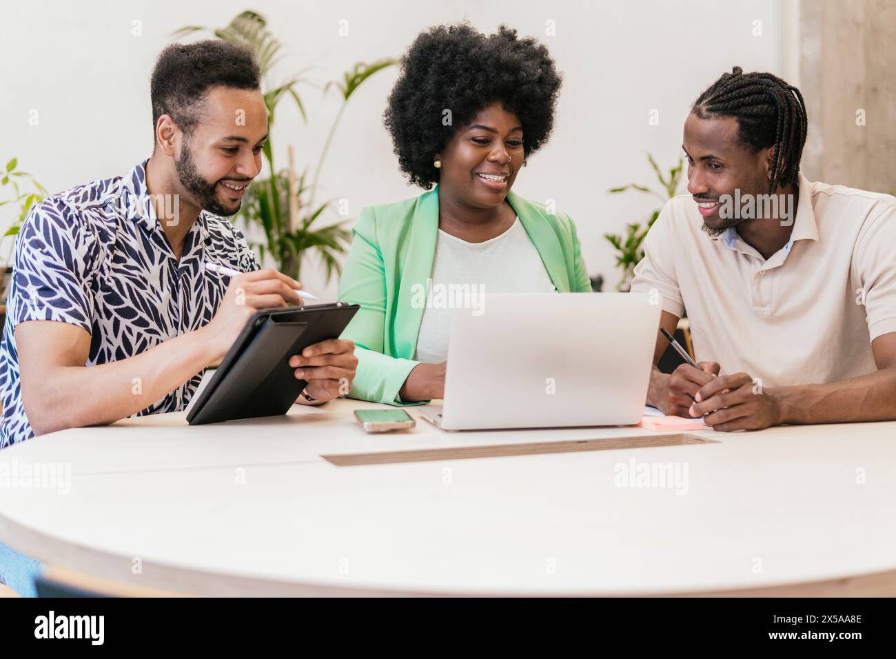 Three professionals engage in a collaborative meeting using a laptop and tablet in a coworking space featuring modern, bright decor Stock Photo