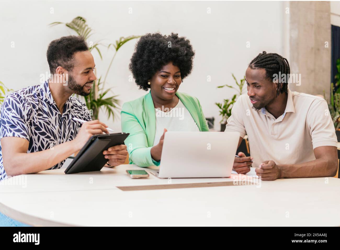 Three professionals engage in collaborative work at a bright coworking space, sharing ideas and resources on a laptop and digital tablet Stock Photo