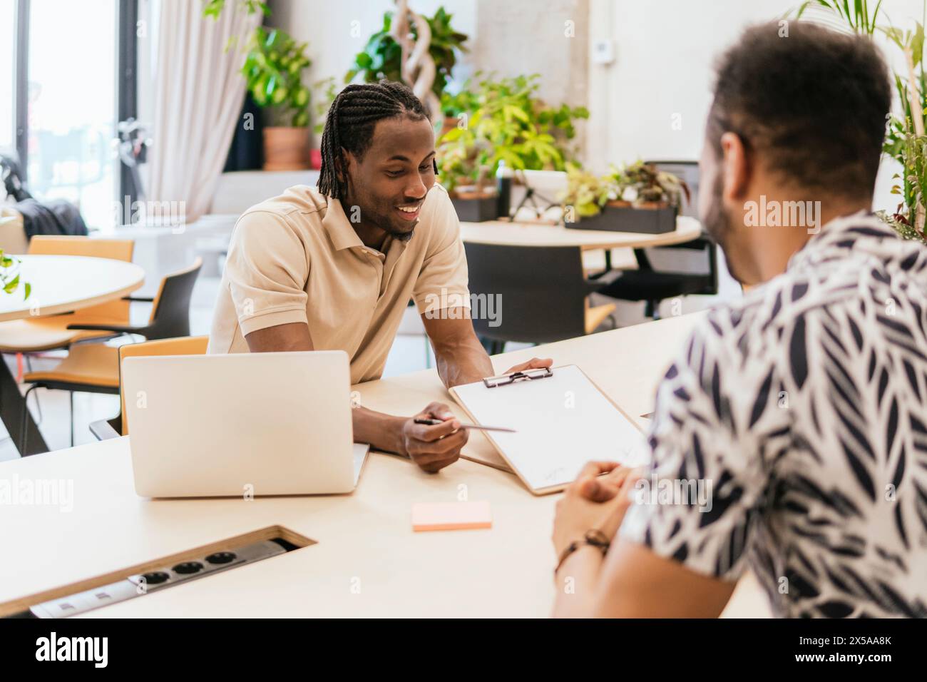 Two professionals engaging in a friendly discussion at a well-lit coworking space, with laptops and note-taking materials at hand Stock Photo