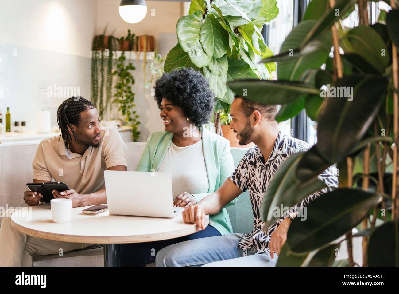 Three professionals collaborate around a laptop in a modern, plant-filled coworking environment, exchanging ideas and enjoying a friendly conversation Stock Photo