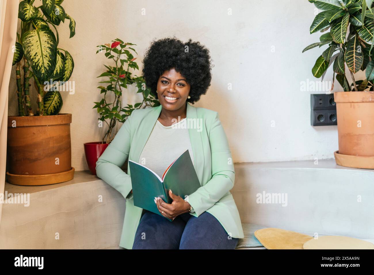 A cheerful woman in a stylish green blazer holds a notebook, seated beside lush indoor plants, radiating confidence and positivity in a coworking spac Stock Photo