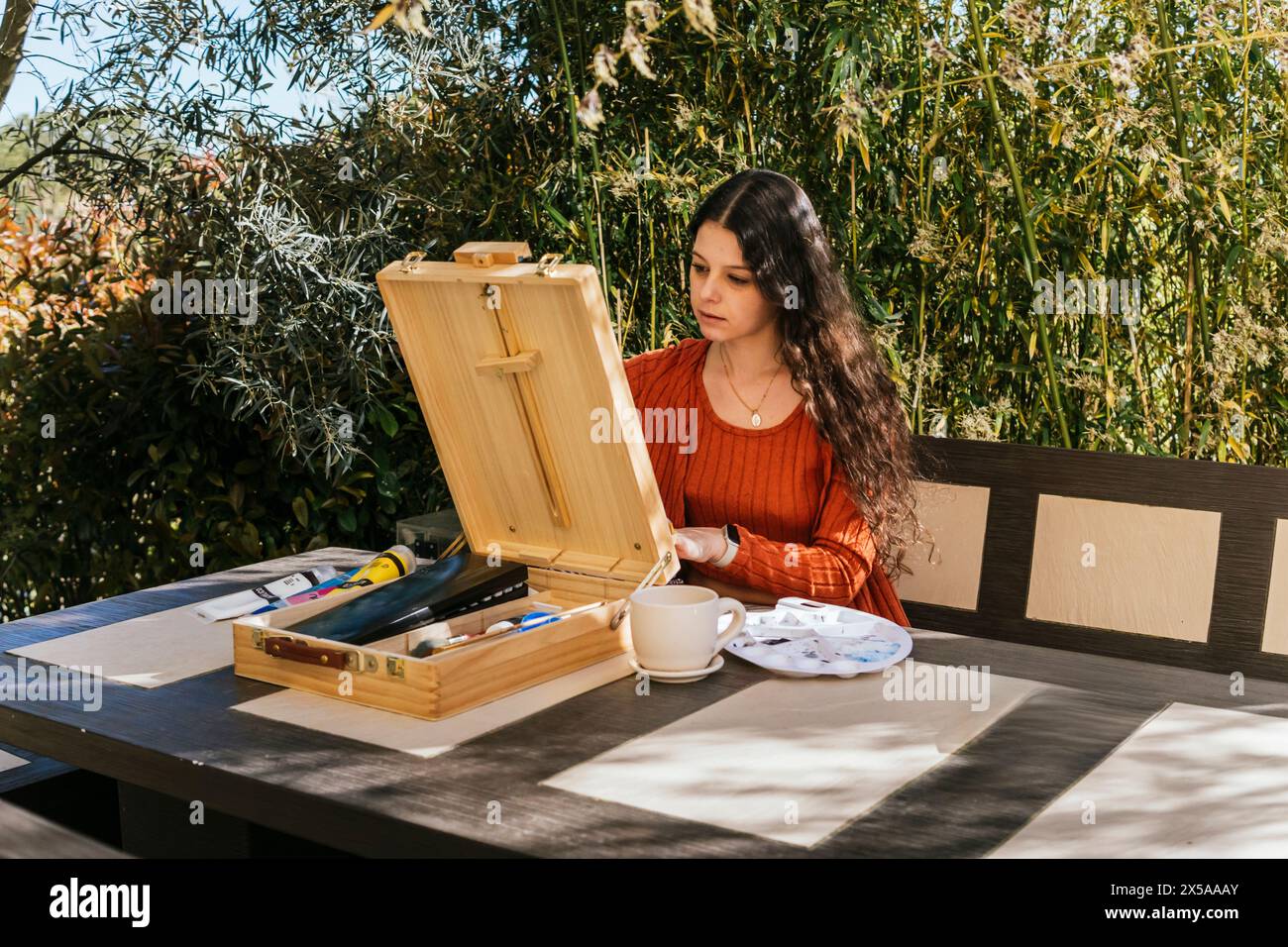 A focused female artist sets up her wooden easel and painting supplies on a sunny patio, surrounded by greenery, ready to start her creative process Stock Photo
