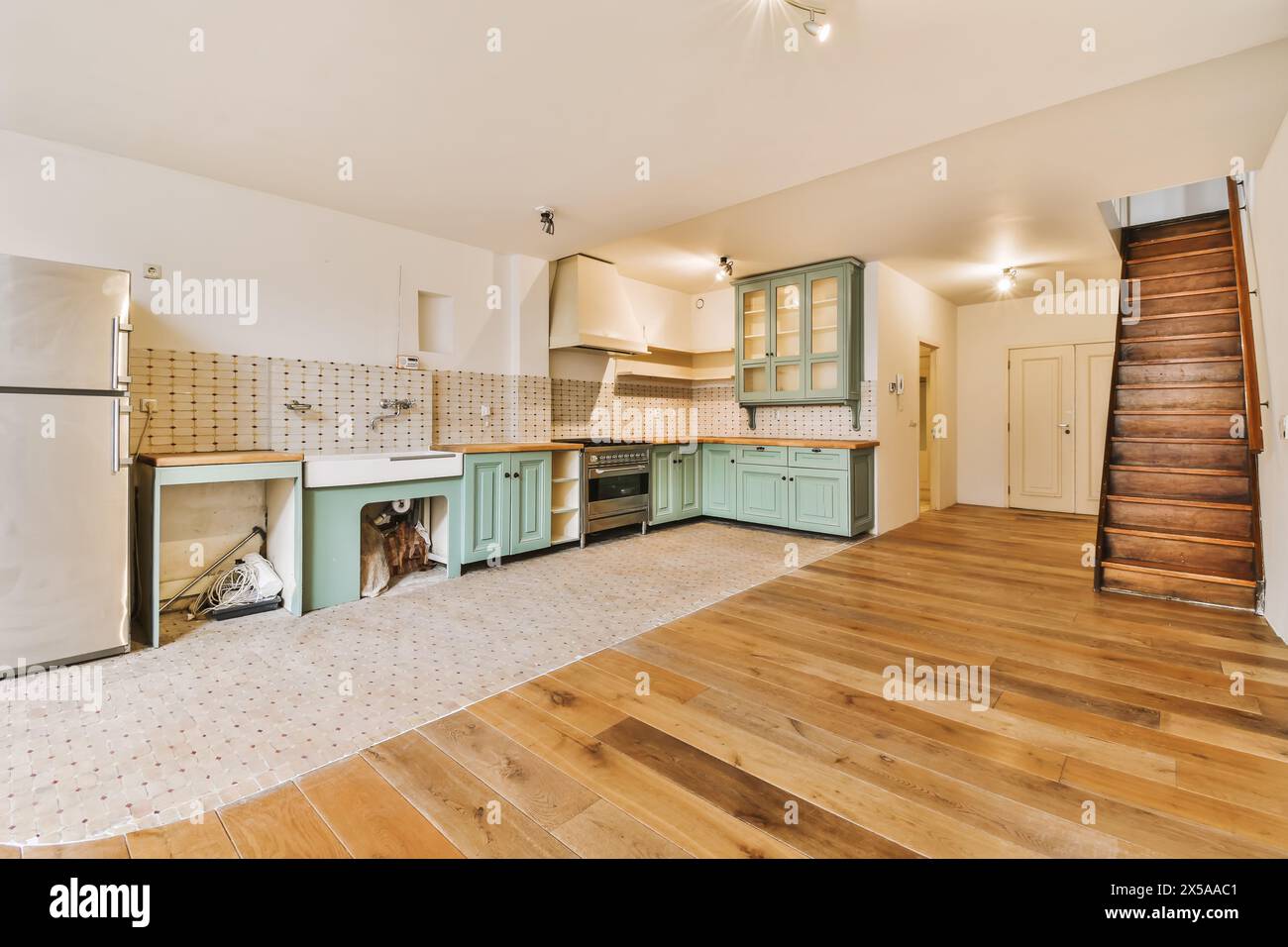 A wide-angle view of a spacious kitchen with mint-colored cabinets, modern appliances, a wooden staircase, and hardwood flooring. Stock Photo
