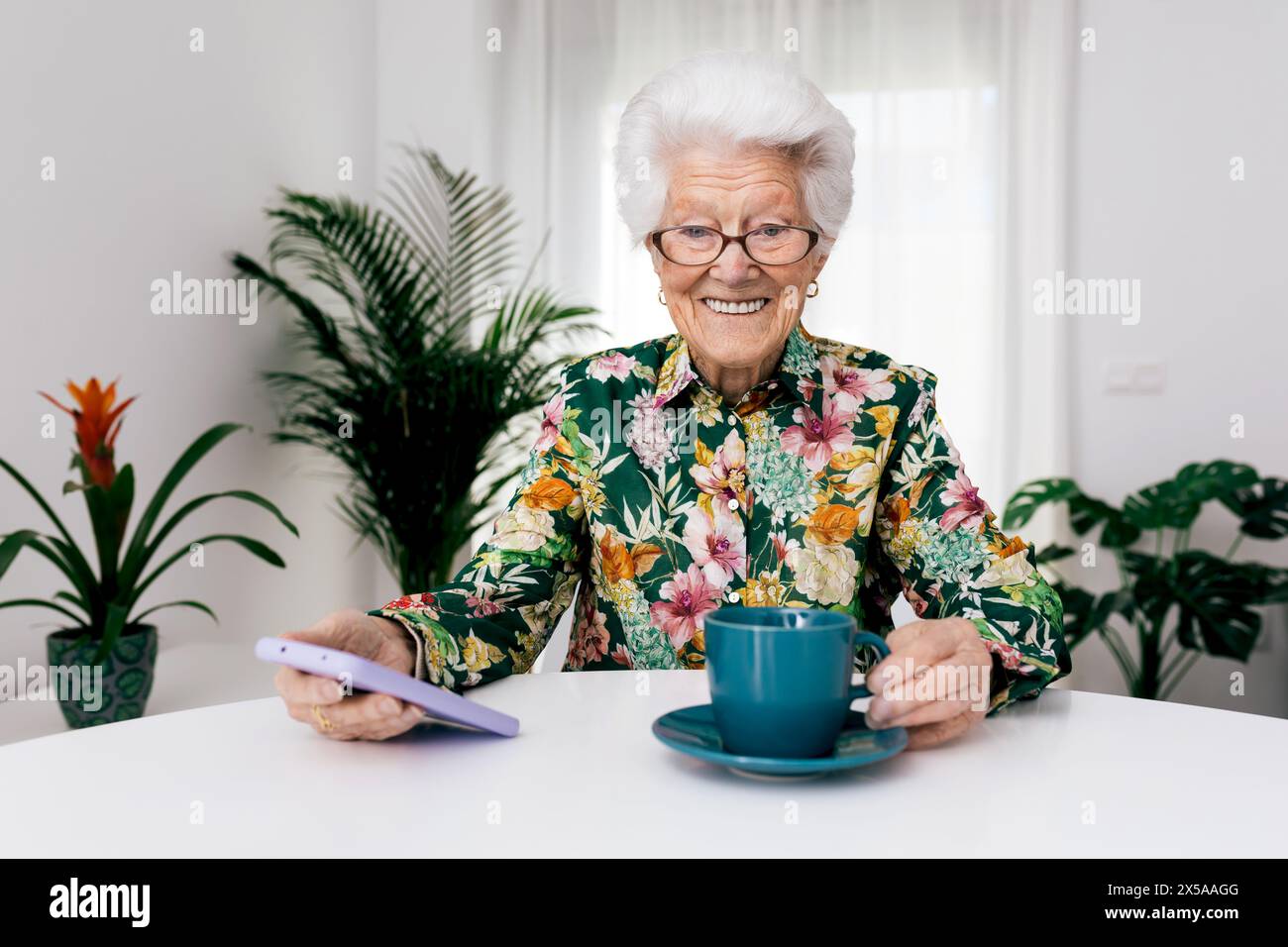 Cheerful senior woman with glasses and a floral shirt enjoying a cup of coffee while holding a smartphone at a white table with houseplants in the bac Stock Photo
