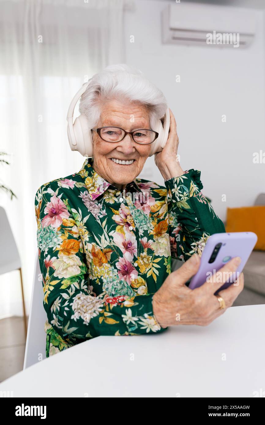 An elderly woman with white hair smiles as she listens to music through white headphones, holding a smartphone in her home Stock Photo