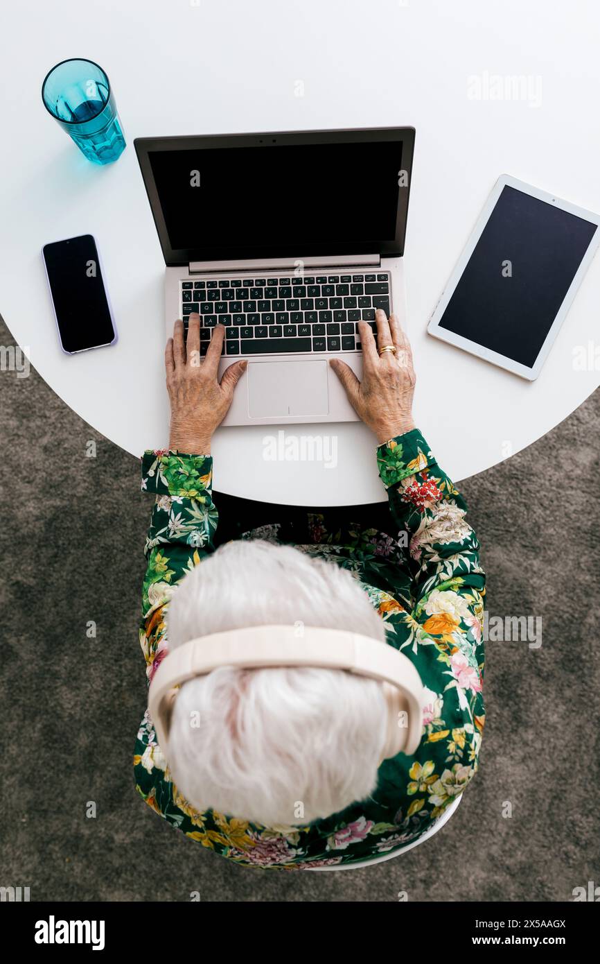 An elderly woman in a floral shirt is seen from above working on her laptop at home, with phone and tablet nearby Stock Photo
