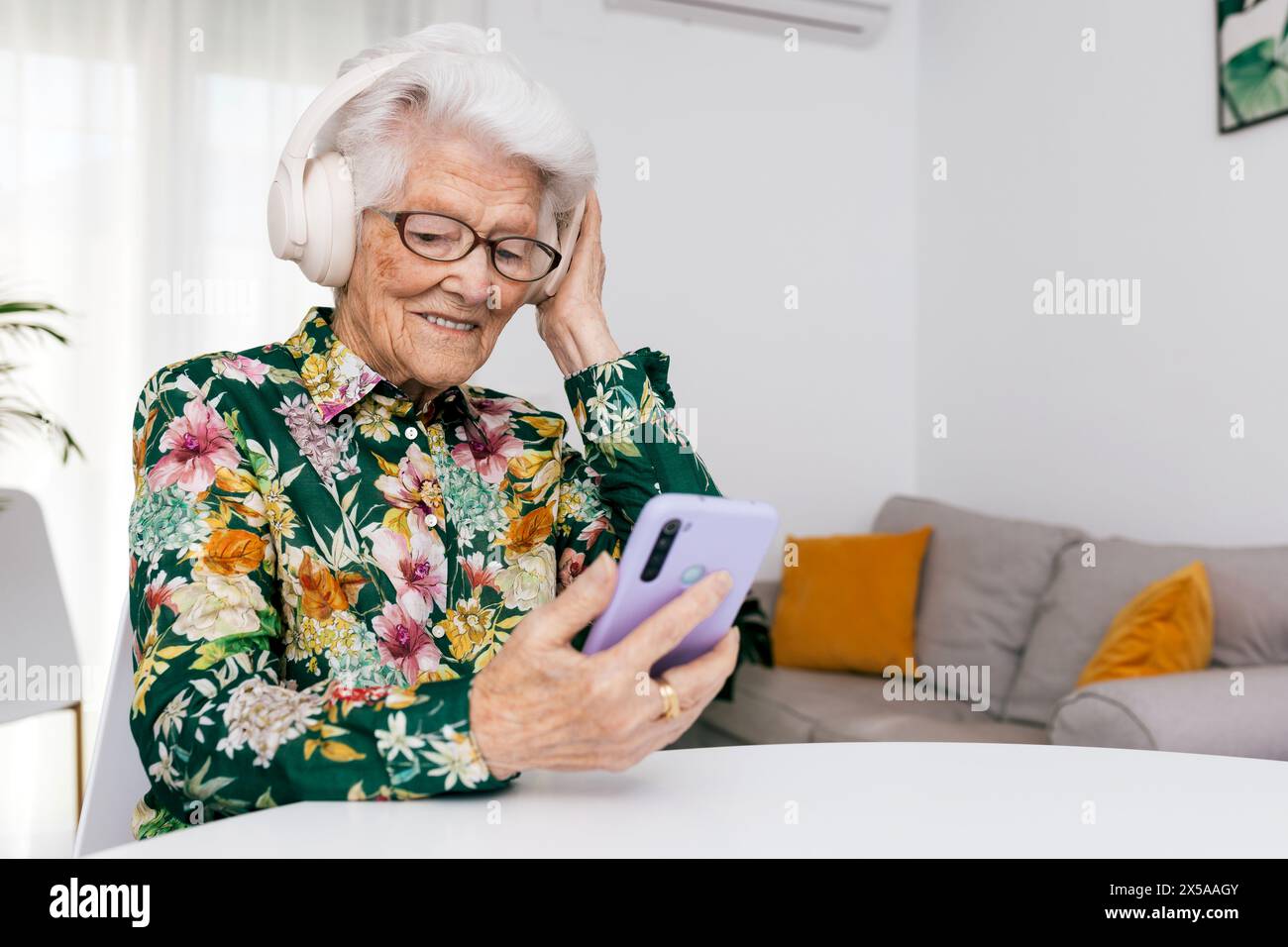 A joyful senior woman with white hair and glasses listens to music using headphones and a smartphone, comfortably seated indoors Stock Photo