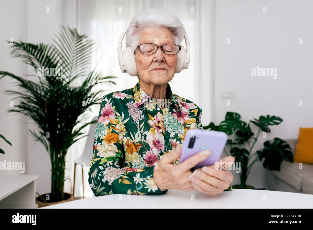 Elderly woman in a floral shirt enjoys music with white headphones while holding a smartphone in a brightly lit living room Stock Photo