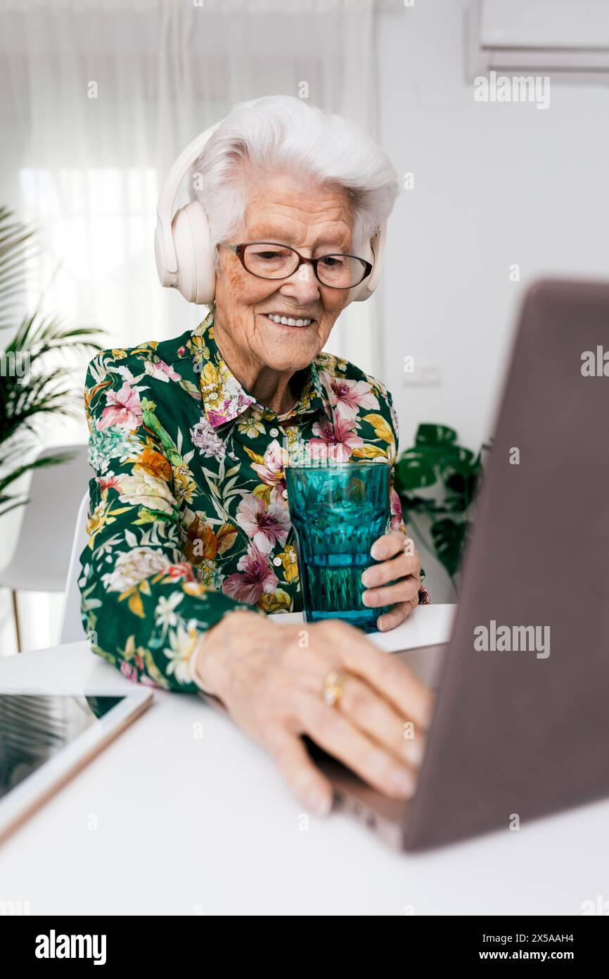 Elderly lady in a floral shirt smiles while using a laptop and wearing headphones, exemplifying active aging and technology use at home Stock Photo